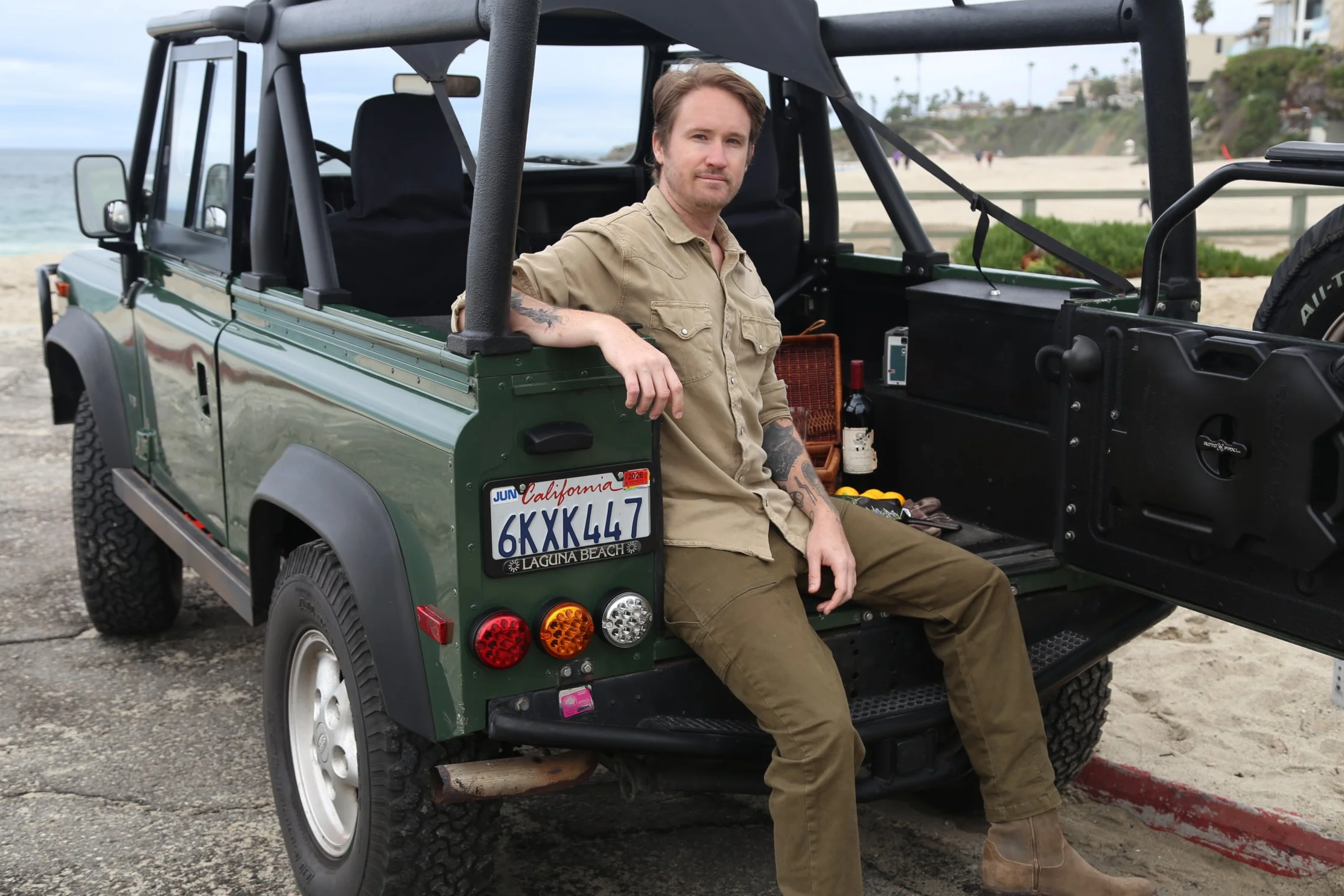 A man sitting in the back of a green Land Rover Defender parked on a beach, with the ocean and houses visible in the background.