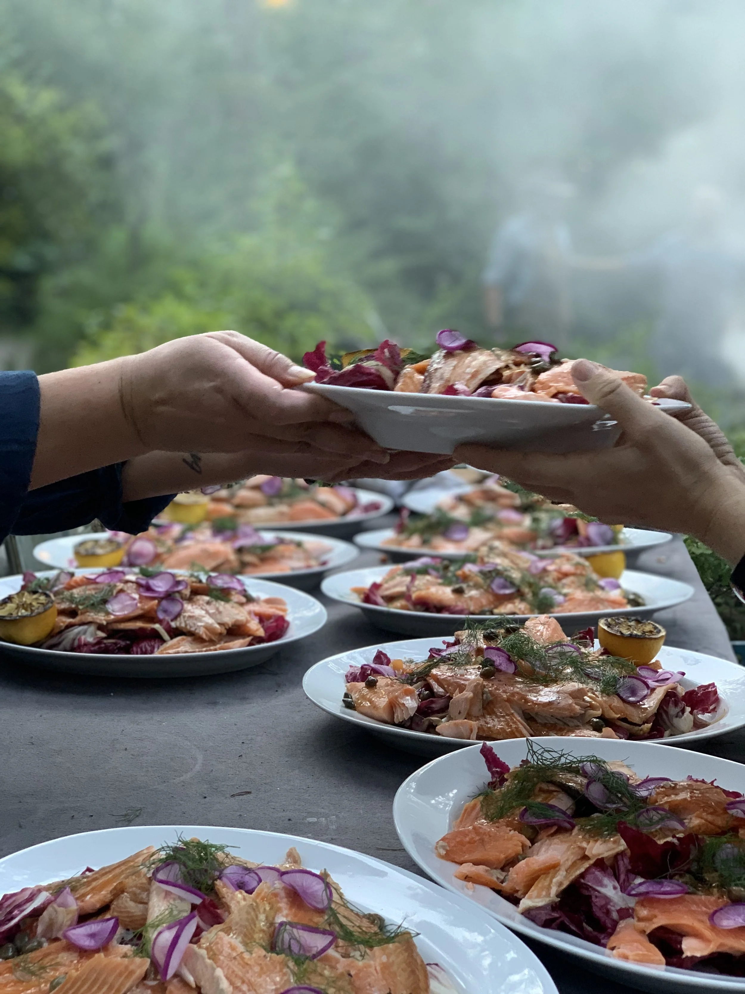 Plates of smoked salmon garnished with sliced red onions, herbs, and other vegetables, arranged on a table at a gathering or event, with a person receiving a plate of food.