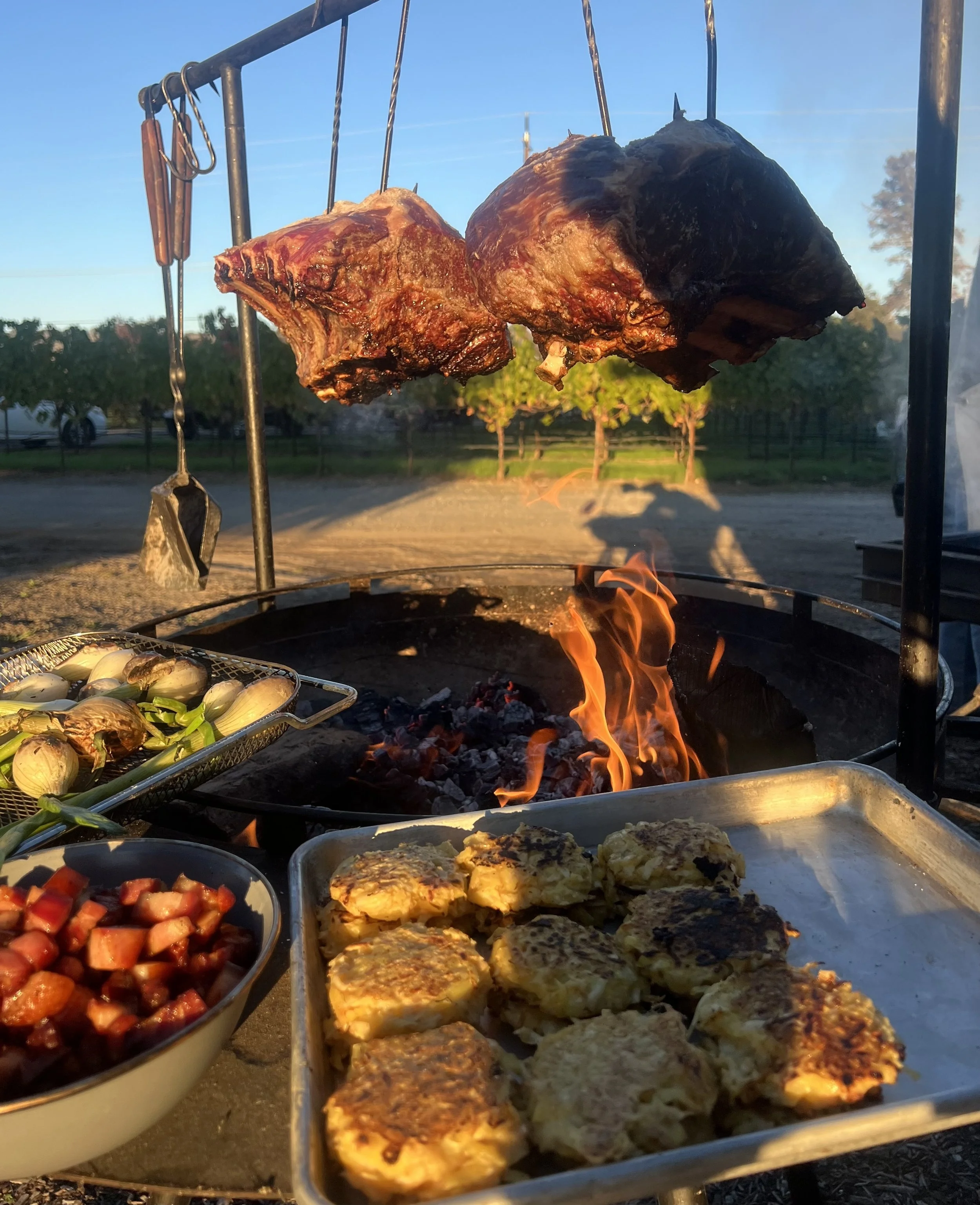 Outdoor barbecue grilling with two large pieces of smoked meat hanging from hooks, surrounded by trays of baked potatoes, grilled vegetables, and diced fruit, with trees and a blue sky in the background.