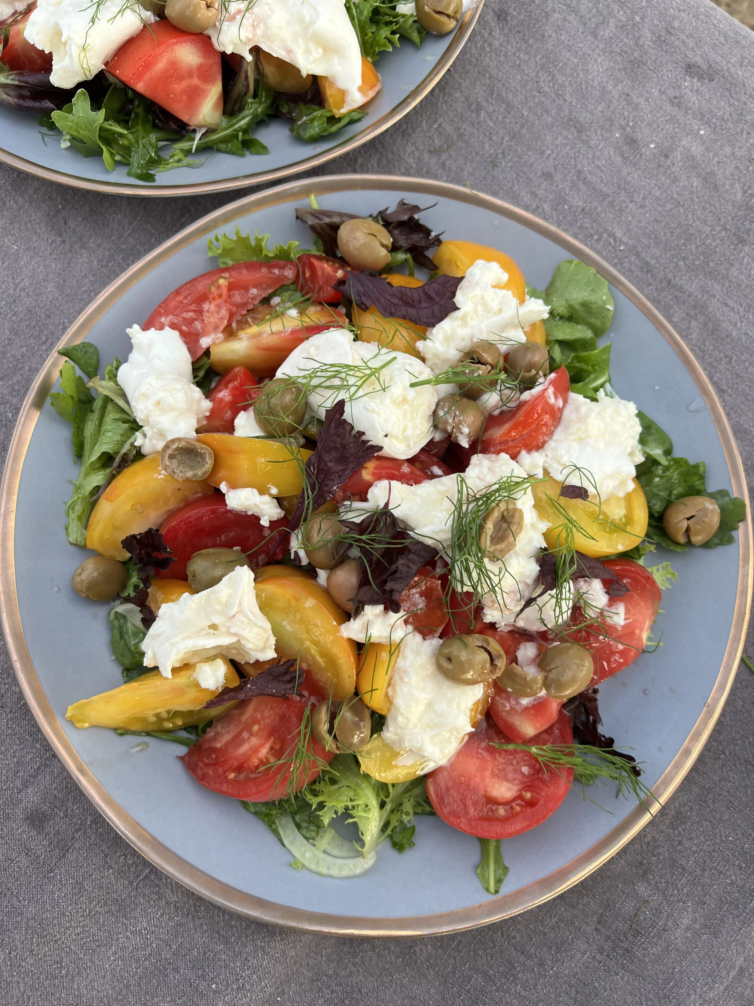 Two plates of salad with tomatoes, mozzarella cheese, yellow and red peppers, green and purple lettuce, green olives, and garnished with fresh herbs on gray table.