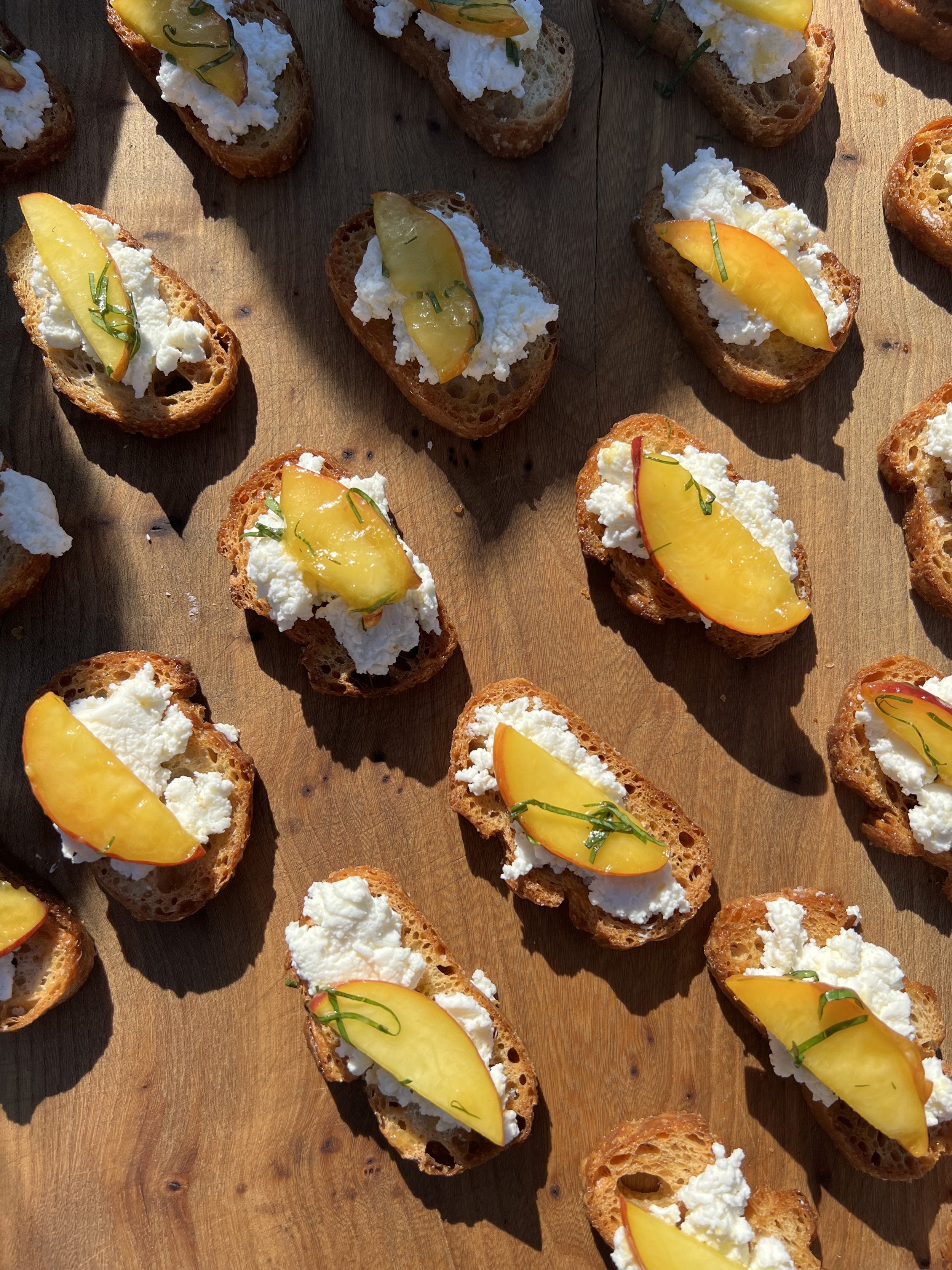 Slices of toasted baguette bread topped with ricotta cheese, yellow peach slices, and chopped chives, arranged on a wooden surface.