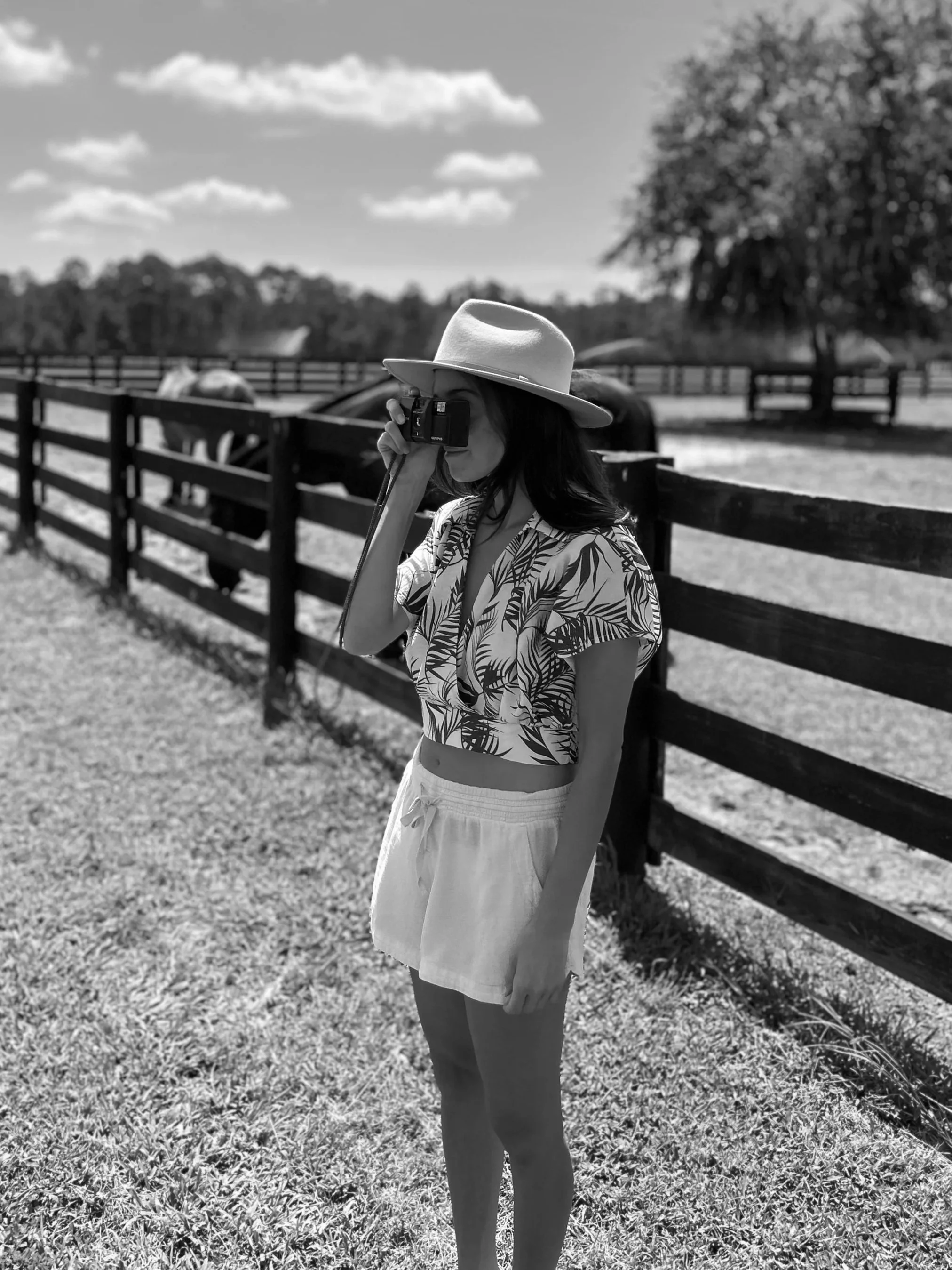 A woman wearing a wide-brimmed hat and patterned shirt standing next to a wooden fence, taking a photo with a camera, outdoors on a sunny day with a tree and horses in the background.