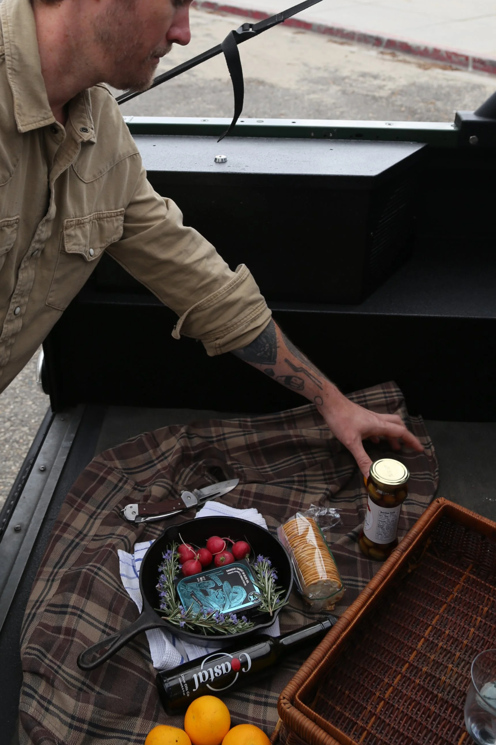 A man setting up a picnic on a plaid blanket with various food items including cherry tomatoes, lemons, a jar of pills, a tin container, and a bottle of Coke. There is a basket and a wine opener on the blanket.