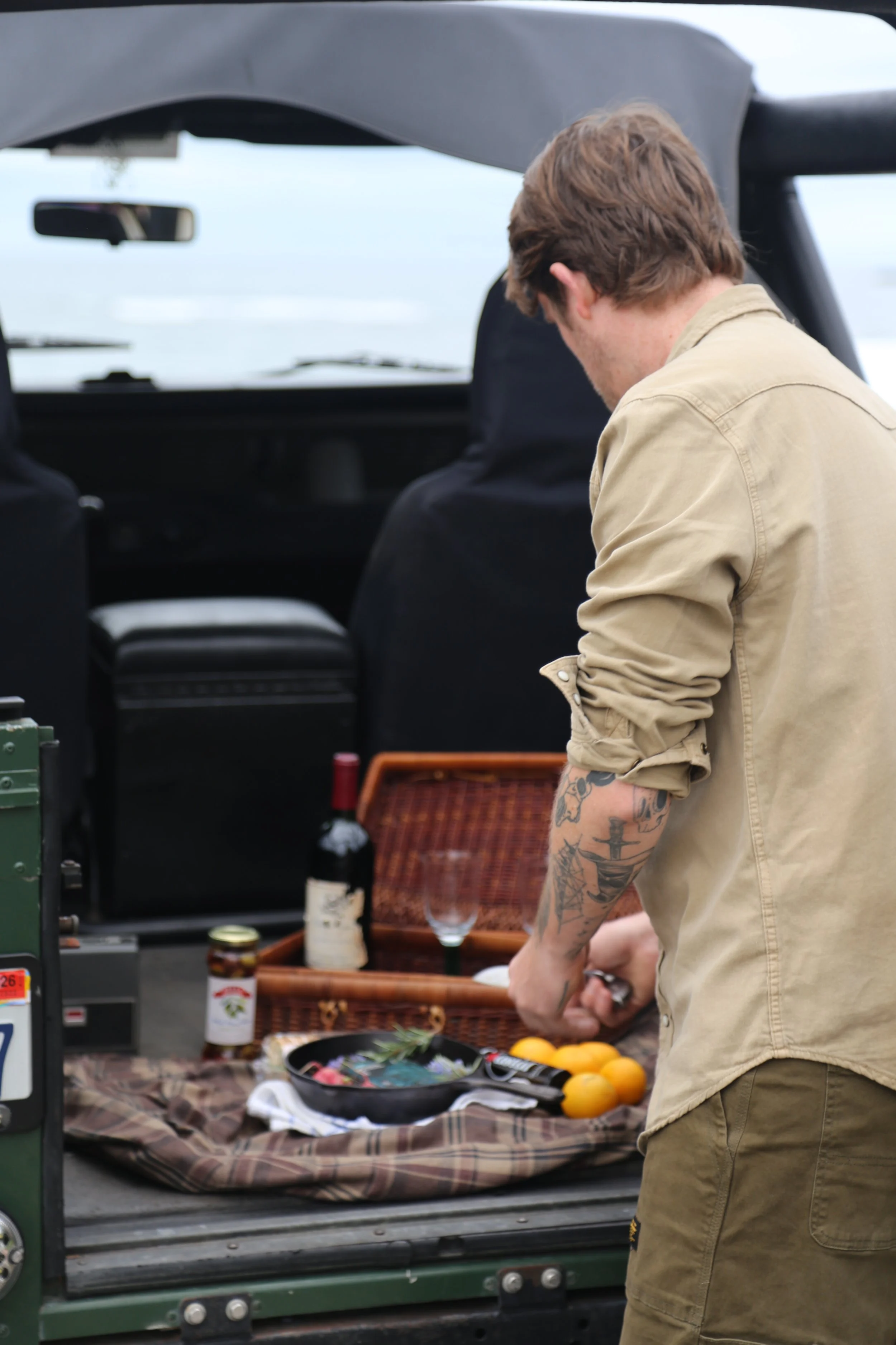A man with tattoos on his forearm standing at an outdoor table with food and drinks, preparing to cook or serve. The table has lemons, jars, and a tray with food, set on a plaid cloth, with a grille or barbecue nearby.