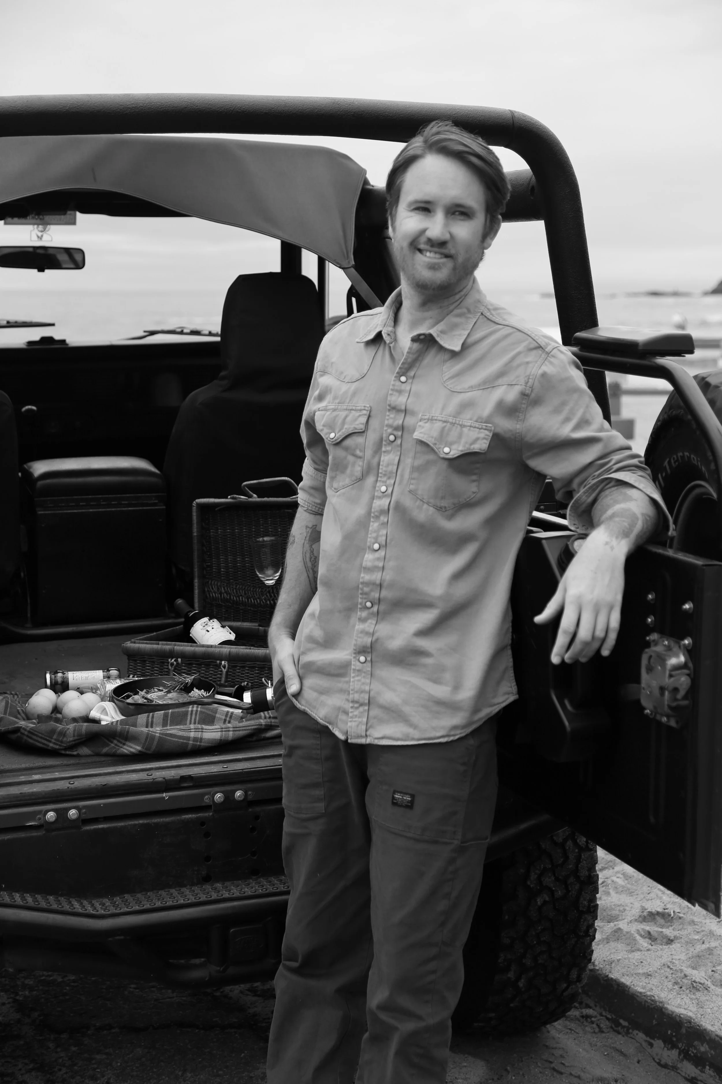 A man stands next to a pickup truck with an open tailgate, at the beach, smiling. The truck bed contains a picnic blanket, eggs, and food items. The man is wearing a long-sleeved button-up shirt and pants.