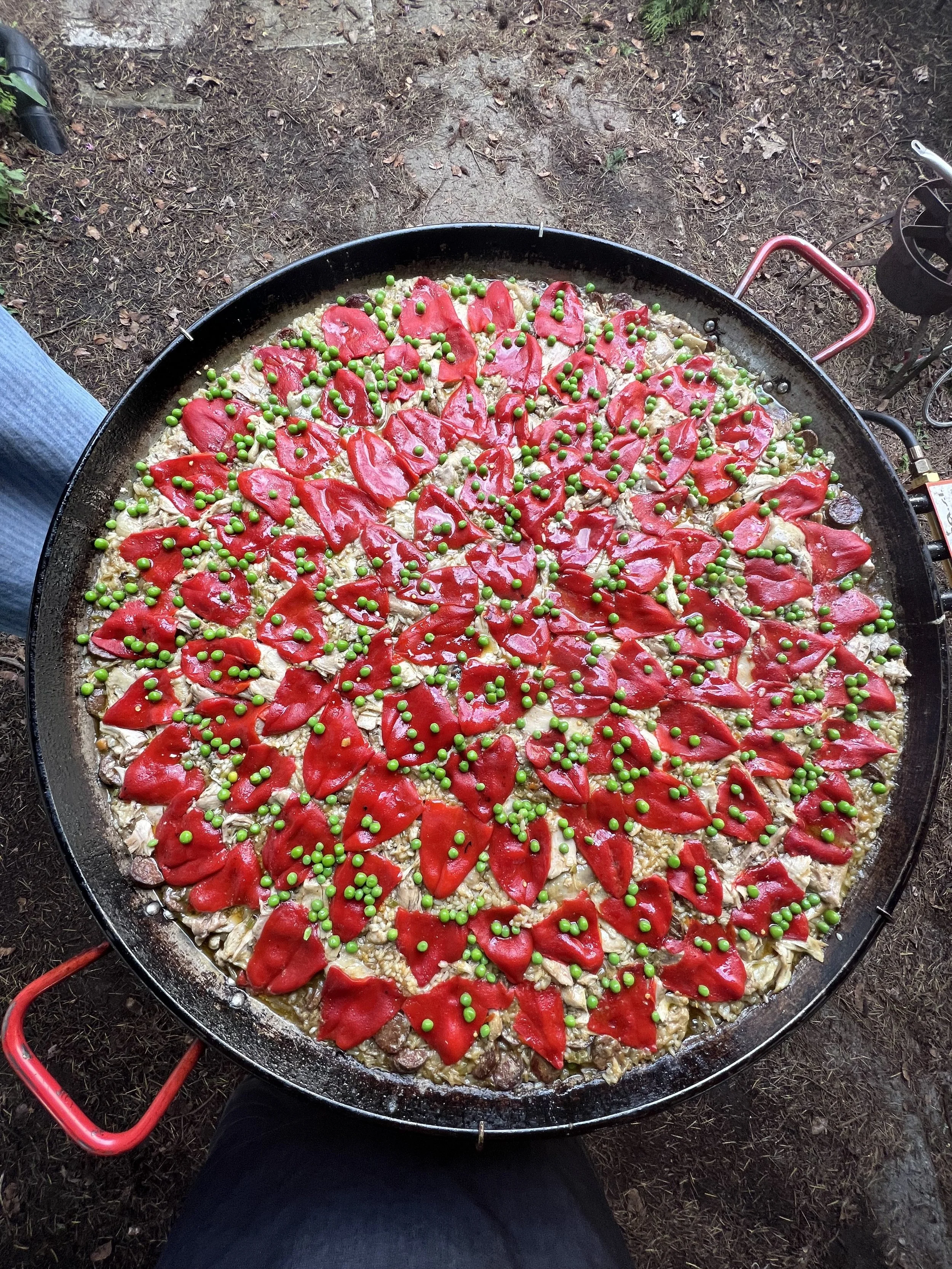 A large outdoor paella cooking pan filled with cooked rice, topped with red bell pepper slices and green peas. The pan is on a portable outdoor stove, with a person's leg visible in the foreground and the ground covered with dirt and leaves surroundi