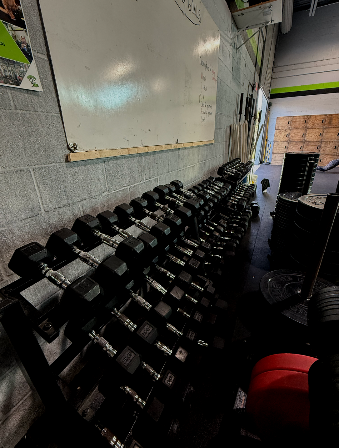A row of black dumbbells on a black metal rack in a gym, with a whiteboard on the wall and plates stacked nearby.