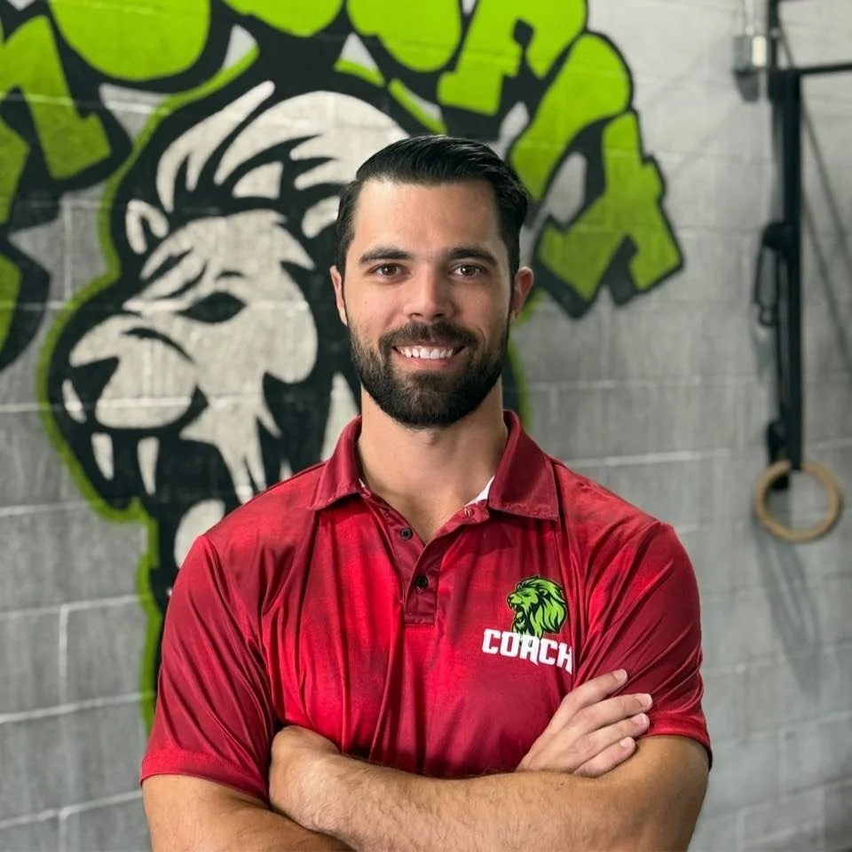 A man with dark hair and a beard, smiling with arms crossed, standing in front of a gym wall with a lion head graffiti logo, wearing a red polo shirt with a green lion logo and the word 'COACH' on it.