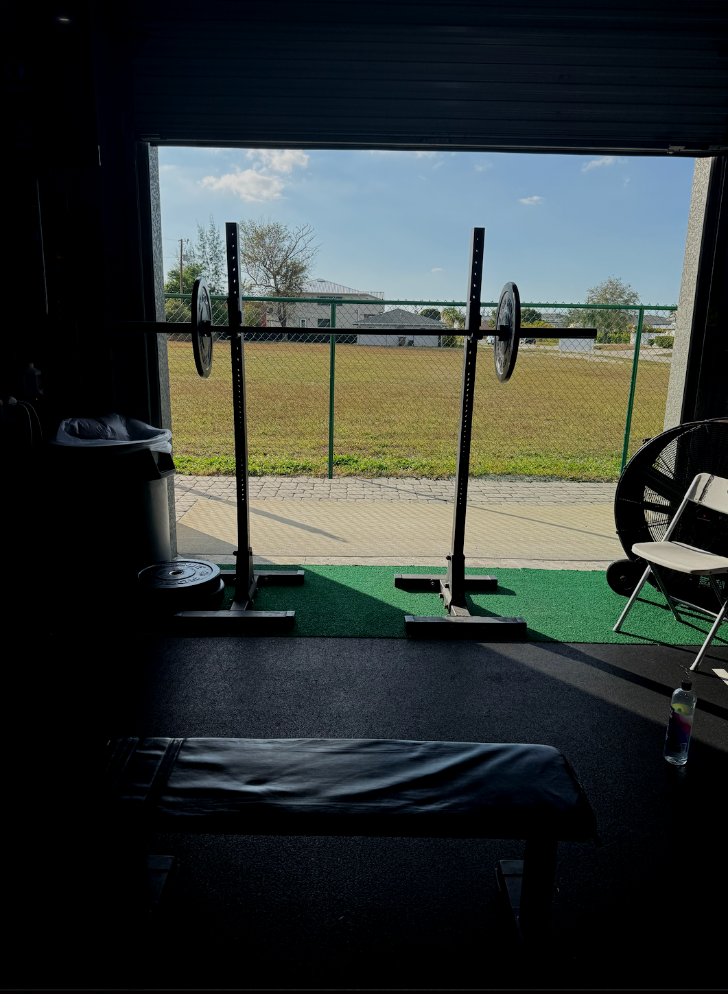 Inside a gym with a squat rack facing open doors, outdoor grass yard, and blue sky