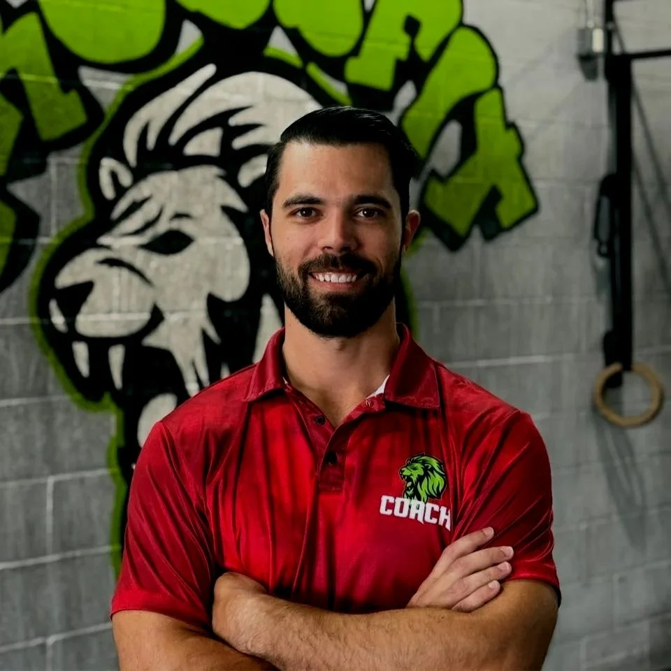 A man with dark hair and a beard wearing a red polo shirt with a green lion logo and the word 'COACH' on it, standing with arms crossed in front of a wall with graffiti of a lion's head in black and white with green outlines.