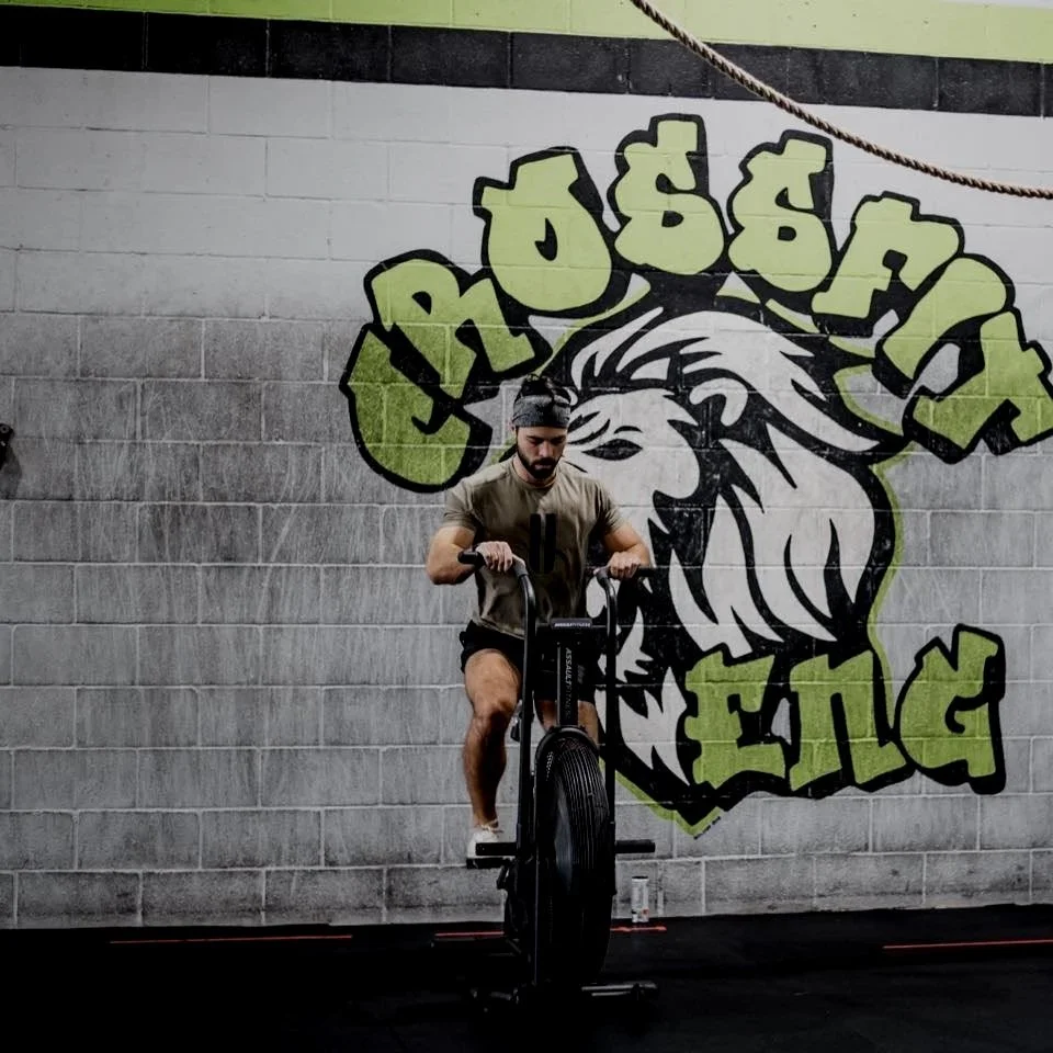 Man riding a stationary exercise bike in a gym with a graffiti-style lion and the words "Rough & Tuff" painted on the wall behind him.