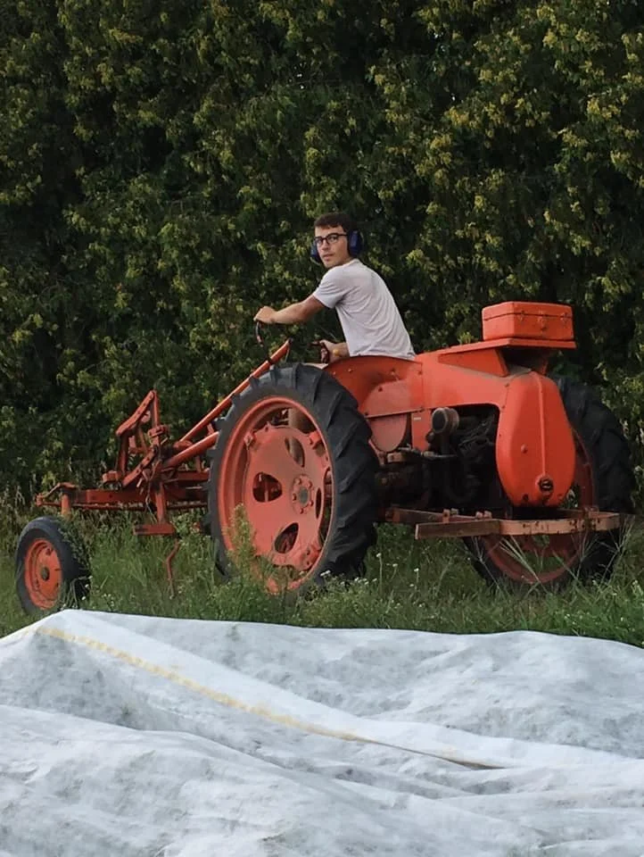A young man wearing glasses and headphones is sitting on an orange vintage tractor in a grassy area with trees behind him.