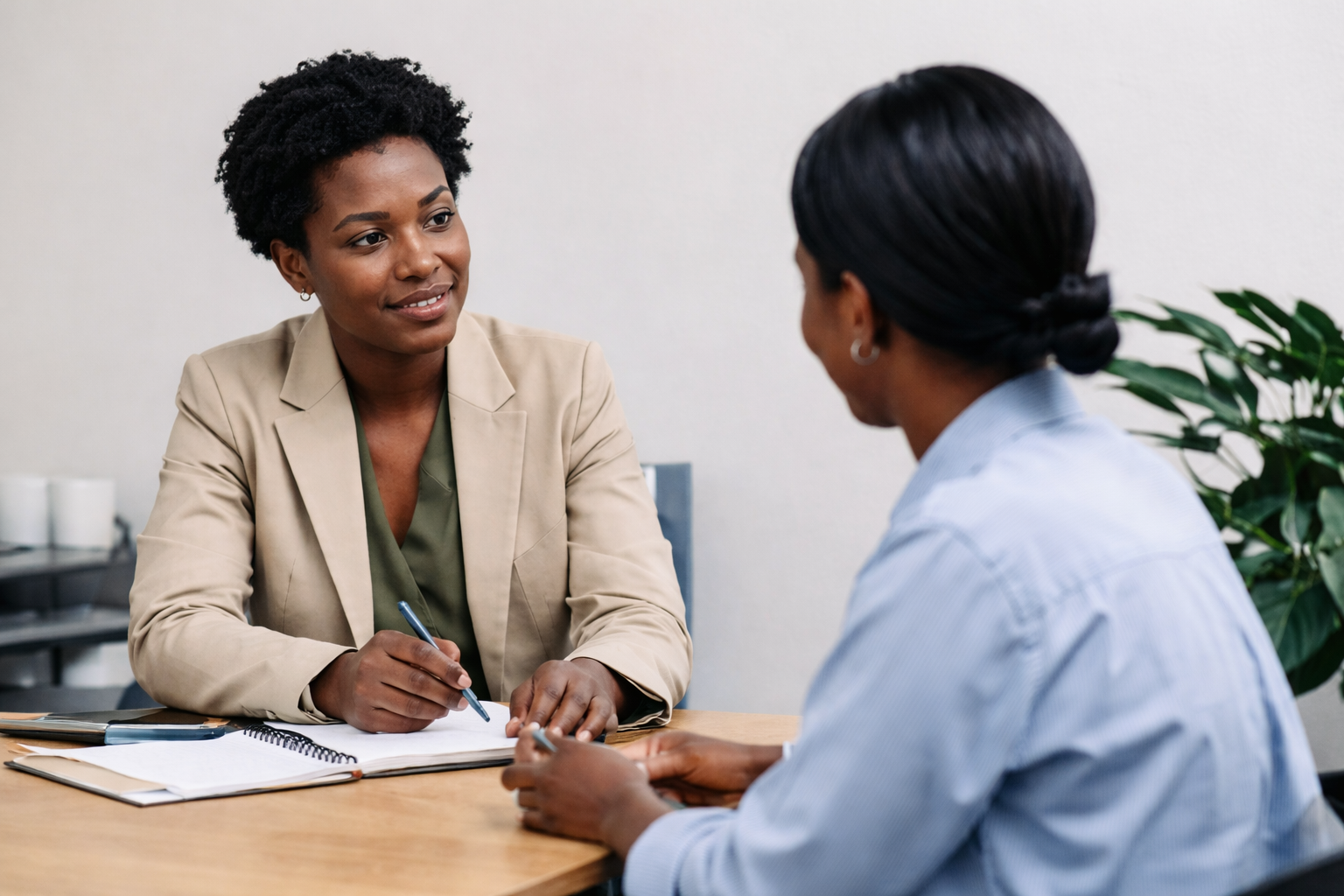 Two women having a conversation at a desk, one with short curly hair and beige blazer, the other with long black hair tied back, wearing a light blue shirt.