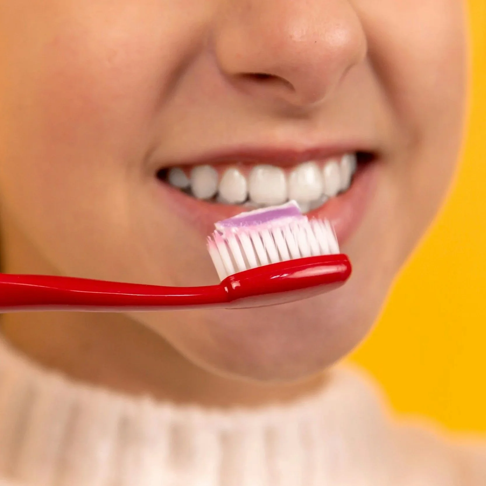 Close-up of a person with a bright smile holding a pink toothbrush with toothpaste near the teeth against a yellow background.