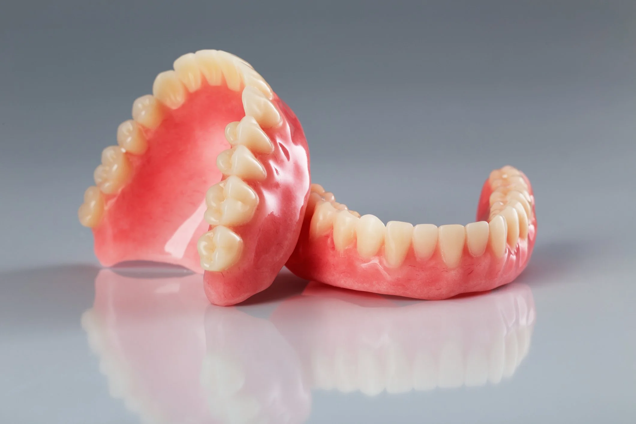 A set of realistic pink and white denture teeth, with one piece upright and the other lying flat on a reflective surface against a gray background.