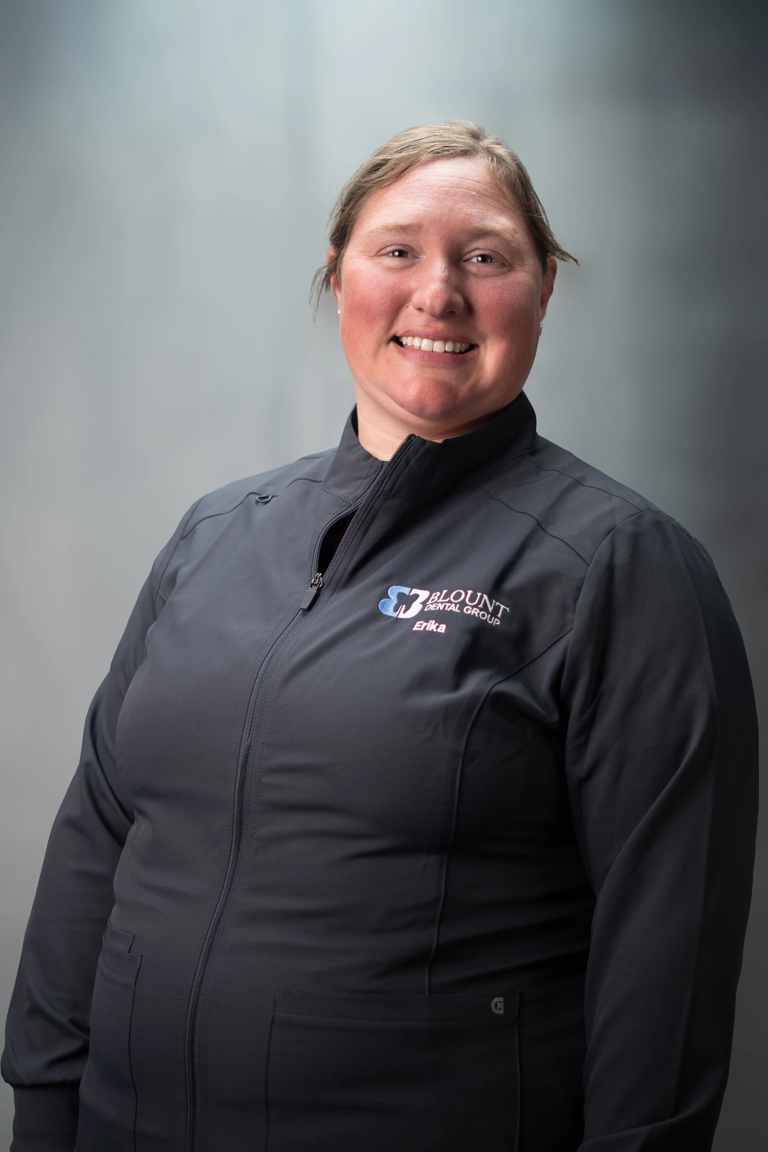 A woman with short hair wearing a black zip-up jacket with the Blount Dental Group logo and her name Erika embroidered on it, smiling against a plain gray background.
