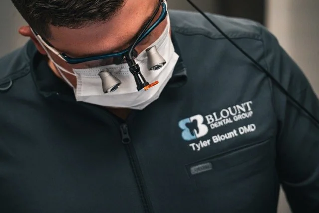 Dental professional wearing magnifying glasses, face mask, and a jacket with 'Blount Dental Group' logo and name tag.