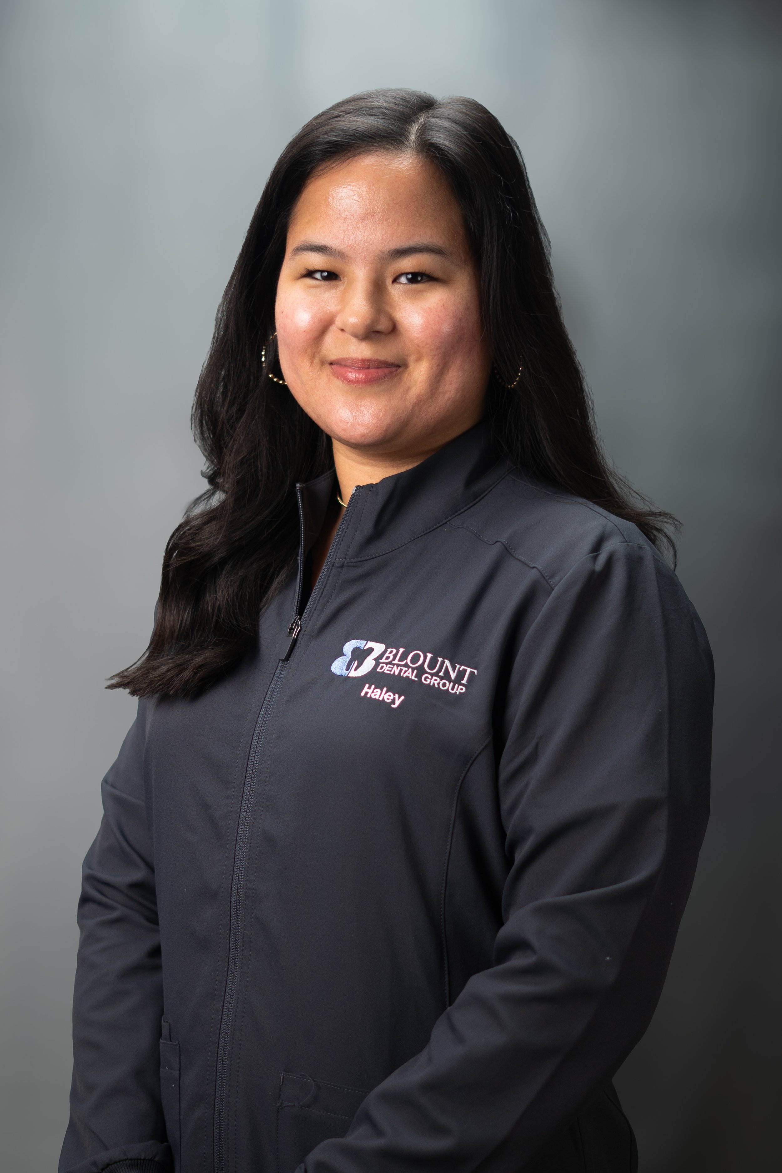 A woman with long dark hair smiling, wearing a black jacket with a logo and the name Haley embroidered on it, standing against a gray background.