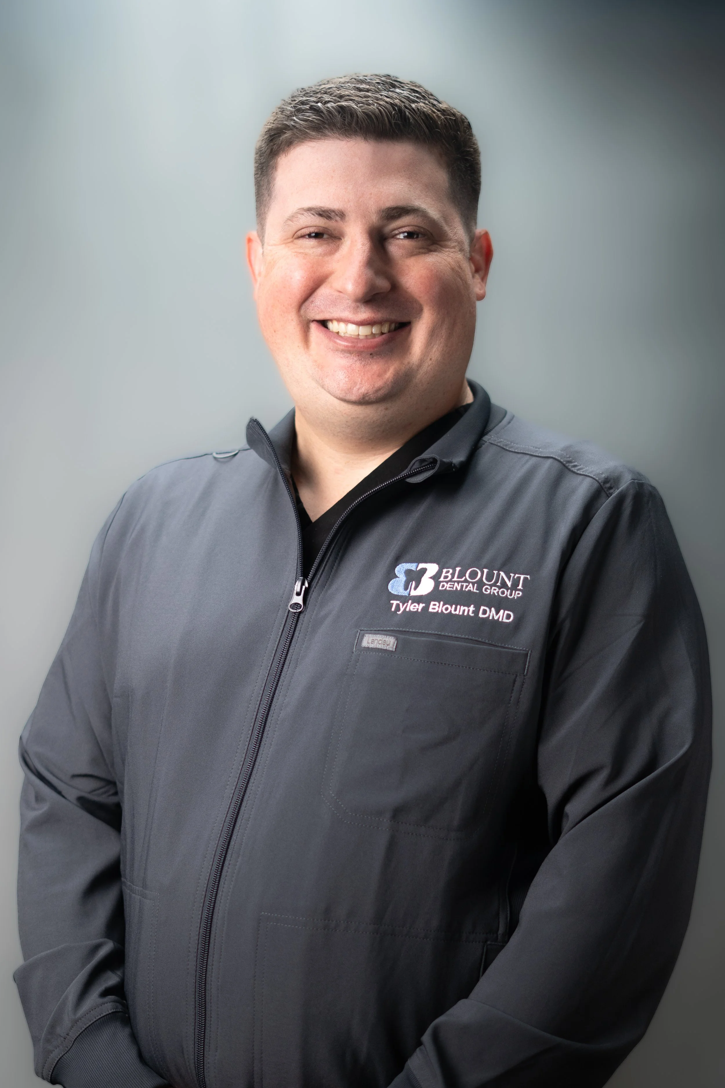 A man smiling, wearing a dark gray blazer with a logo and name tag that reads 'Blount Dental Group, Tyler Blount DMD' against a plain gray background.