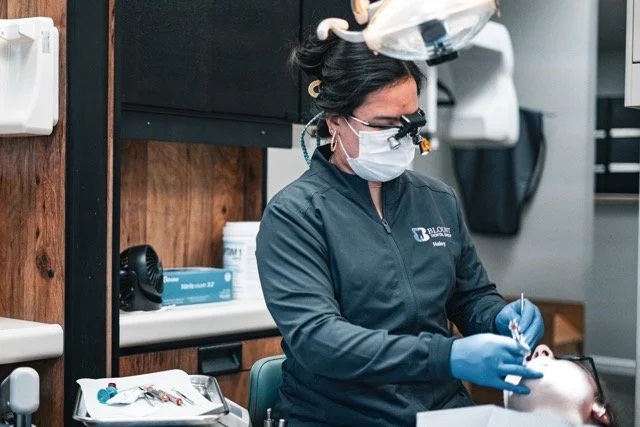 Dental professional wearing a mask, magnifying glasses, and gloves working on a patient in a dental office.