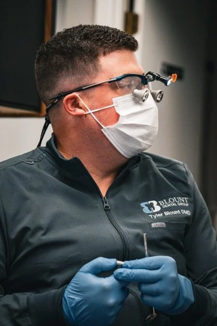 A dentist wearing magnifying loupes, a face mask, and blue gloves standing in a clinical setting.