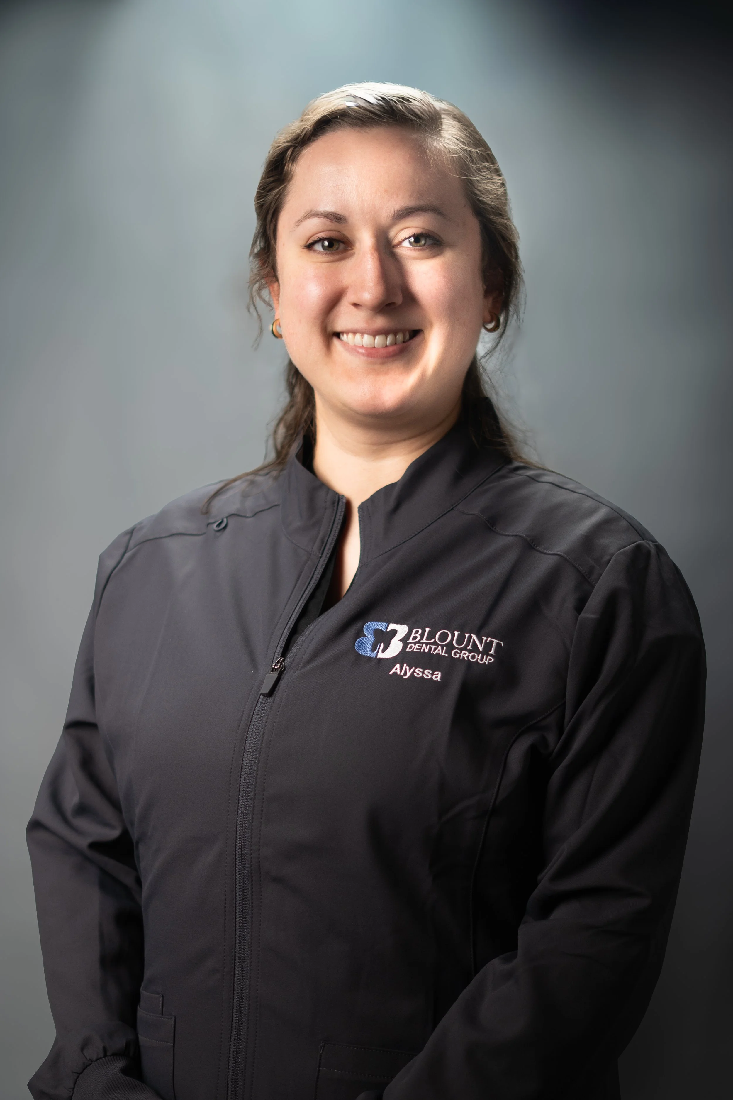 A woman with brown hair smiling, wearing a black jacket with a logo for Blount Dental Group and the name Alyssa embroidered on it, standing against a gray background.