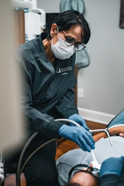 Dental professional in scrubs, face mask, and gloves performing a dental procedure on a patient in a clinic setting.