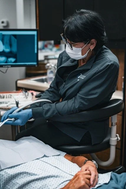 Healthcare professional wearing a mask and gloves attending to a patient in a medical setting with monitors and medical equipment in the background.