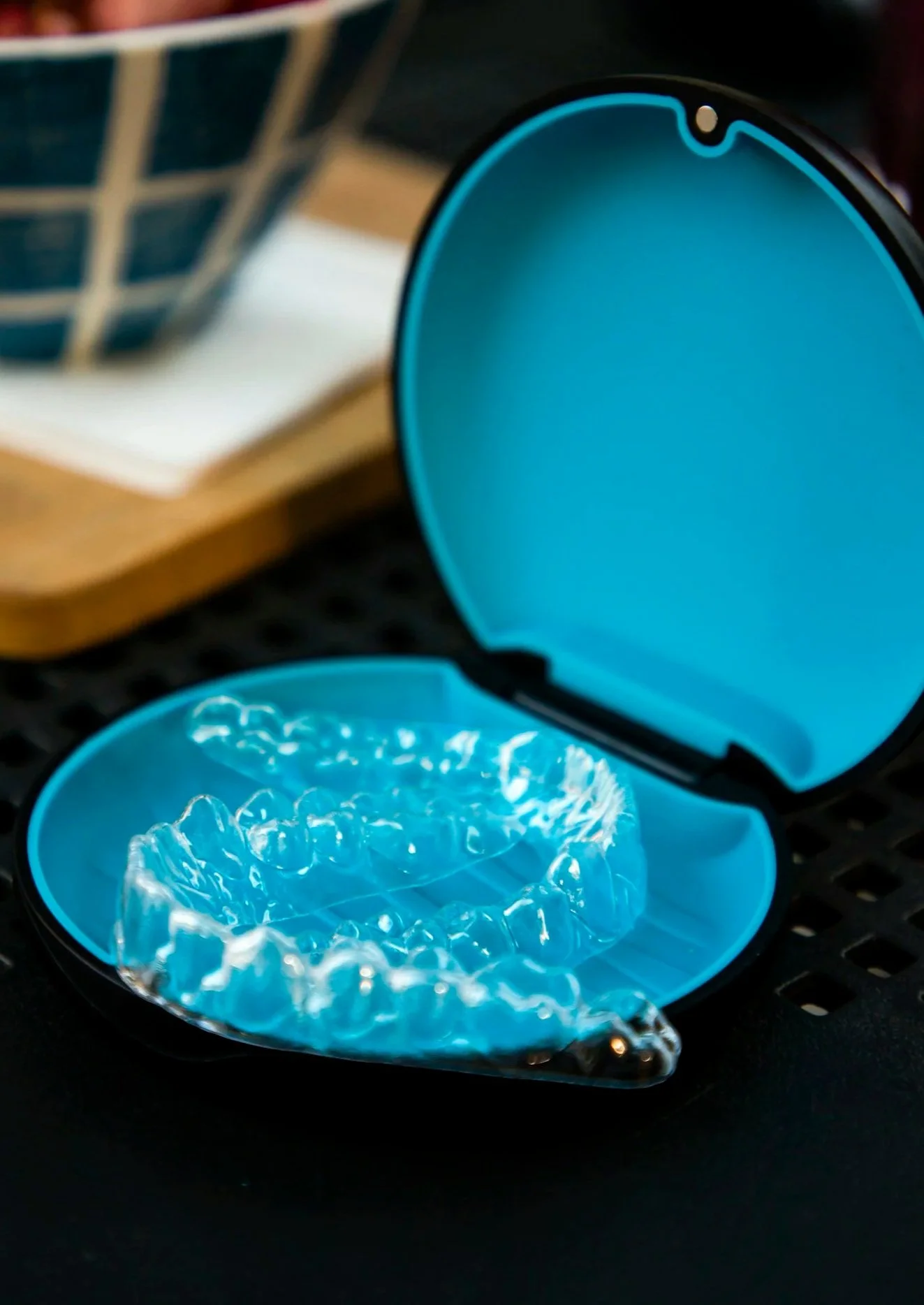 Clear dental aligner inside a blue case, placed on a dark surface with a blurred book or container in the background.
