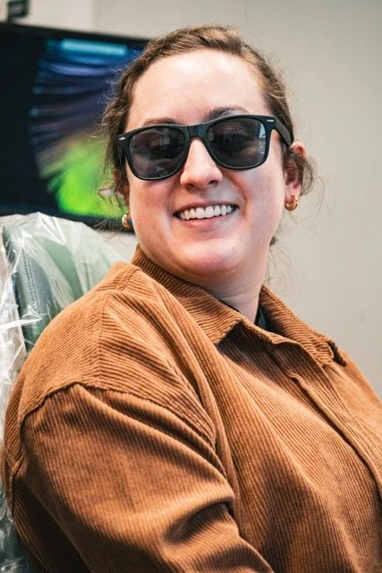 Young woman wearing sunglasses smiling, sitting in a chair with a light brown corduroy jacket in an indoor setting.