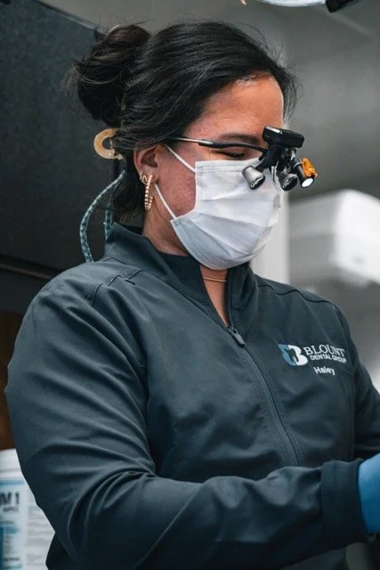 A woman wearing protective glasses with a headlamp, a face mask, and a navy blue jacket with a dental logo, working in a dental clinic.