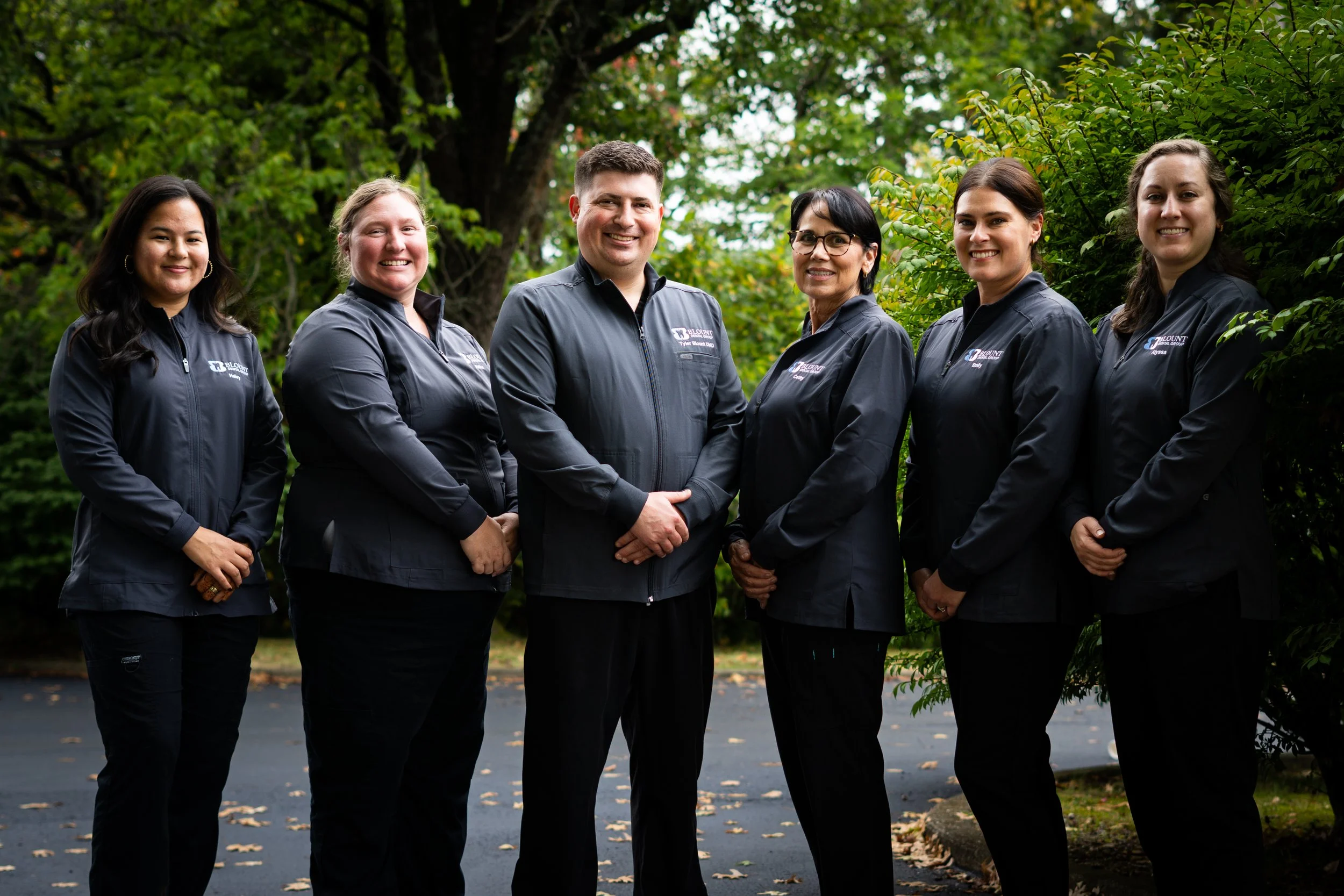 Group of six professional staff members standing outdoors, wearing matching dark jackets with a logo, in a lush green park with trees and fallen leaves.