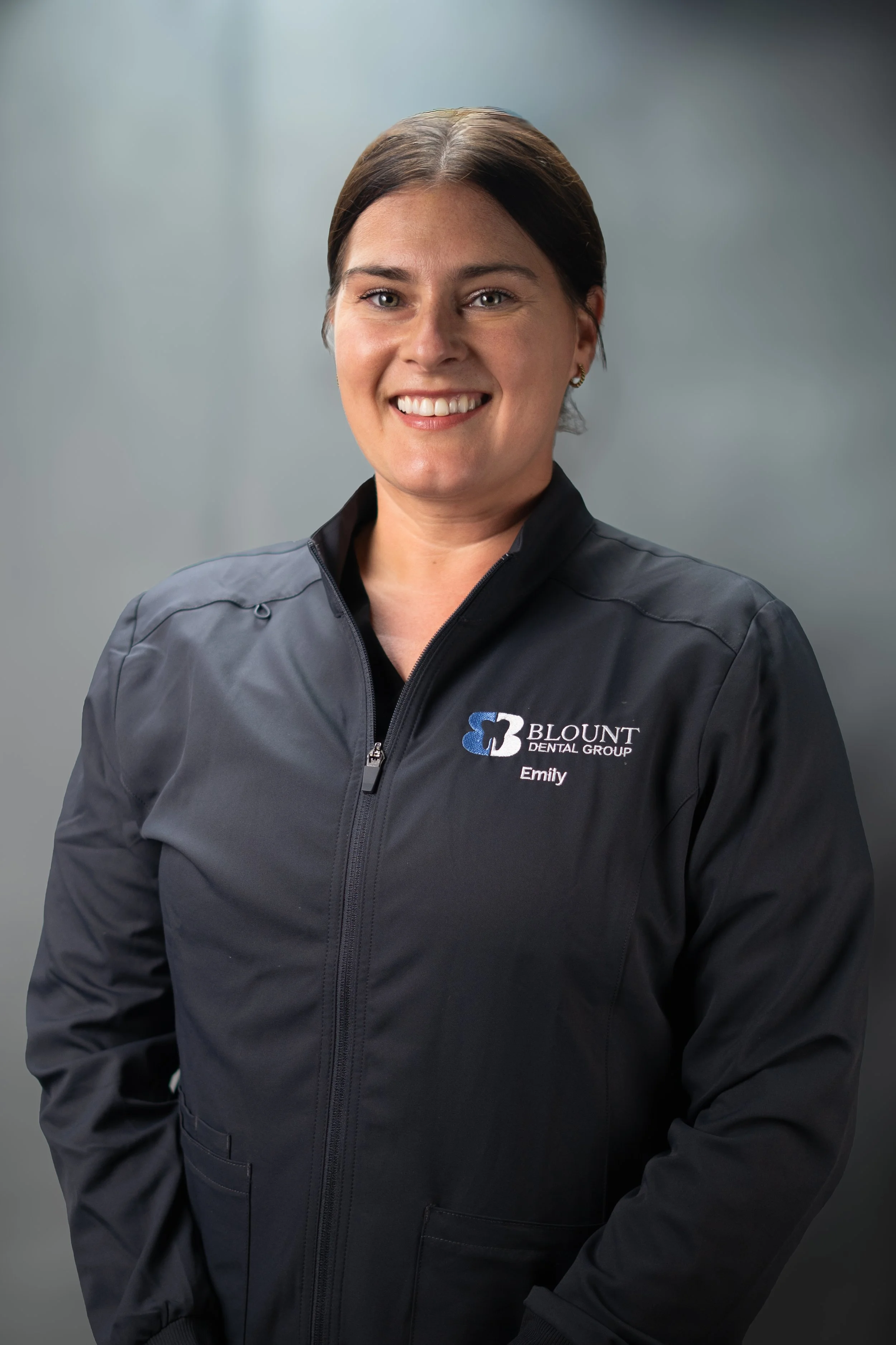 Professional portrait of a woman with brown hair in a black blazer and jacket, smiling, with a blurred gray background.