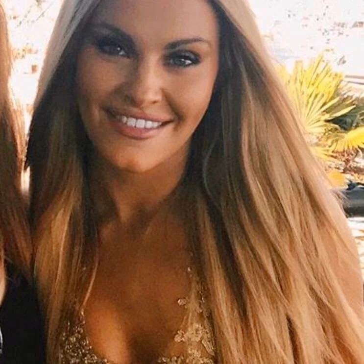 A smiling woman with long, light brown hair, wearing a gold top with sequins, standing outdoors with a blurred background of trees and bright sky.