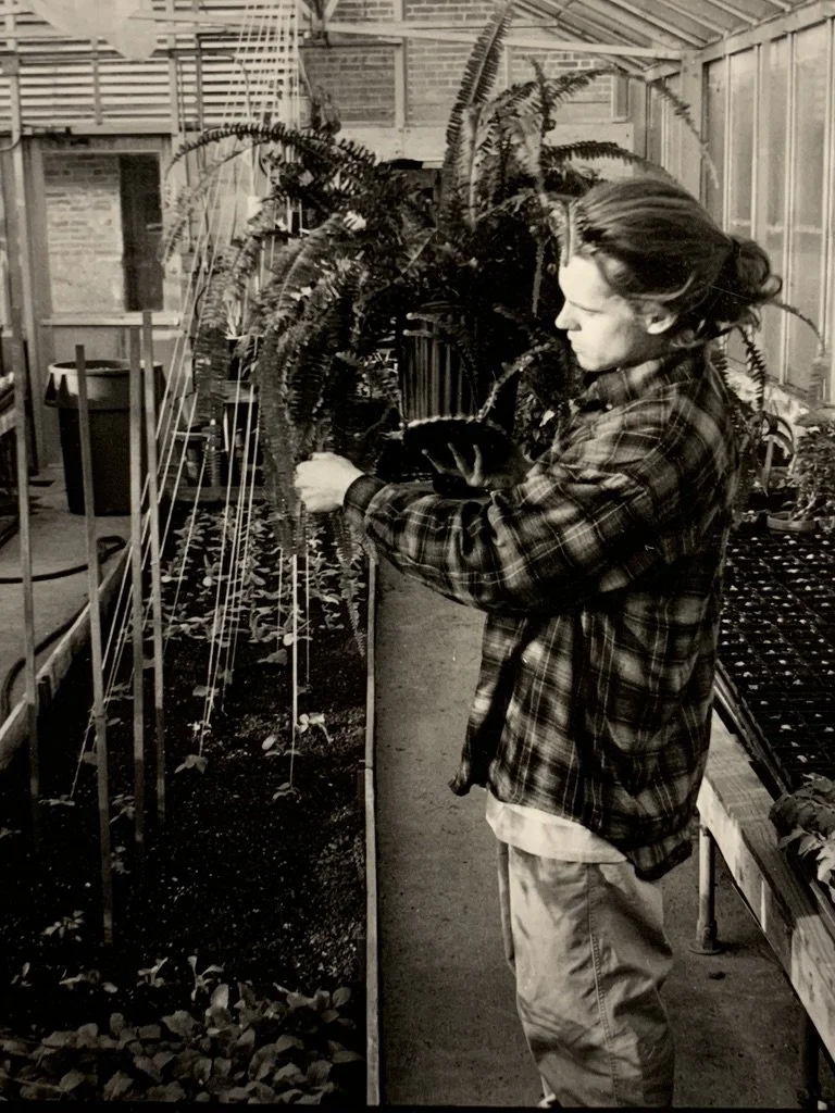 Ryan Douglas in a plaid jacket and pants in a greenhouse, examining plants. There are various plants, including ferns, and gardening tables around.