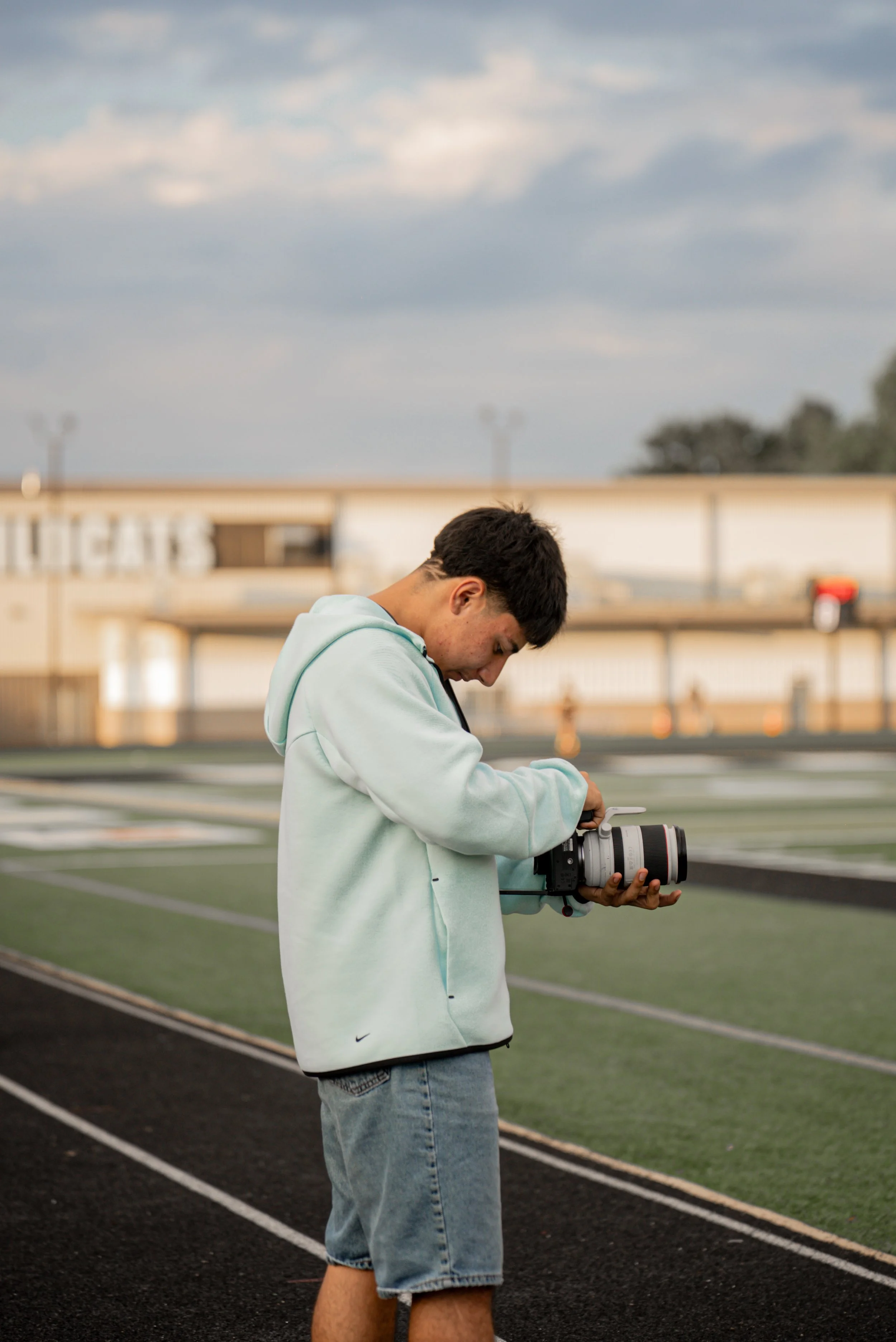 A young man wearing a light blue hoodie and denim shorts standing on a track field, examining a professional camera.
