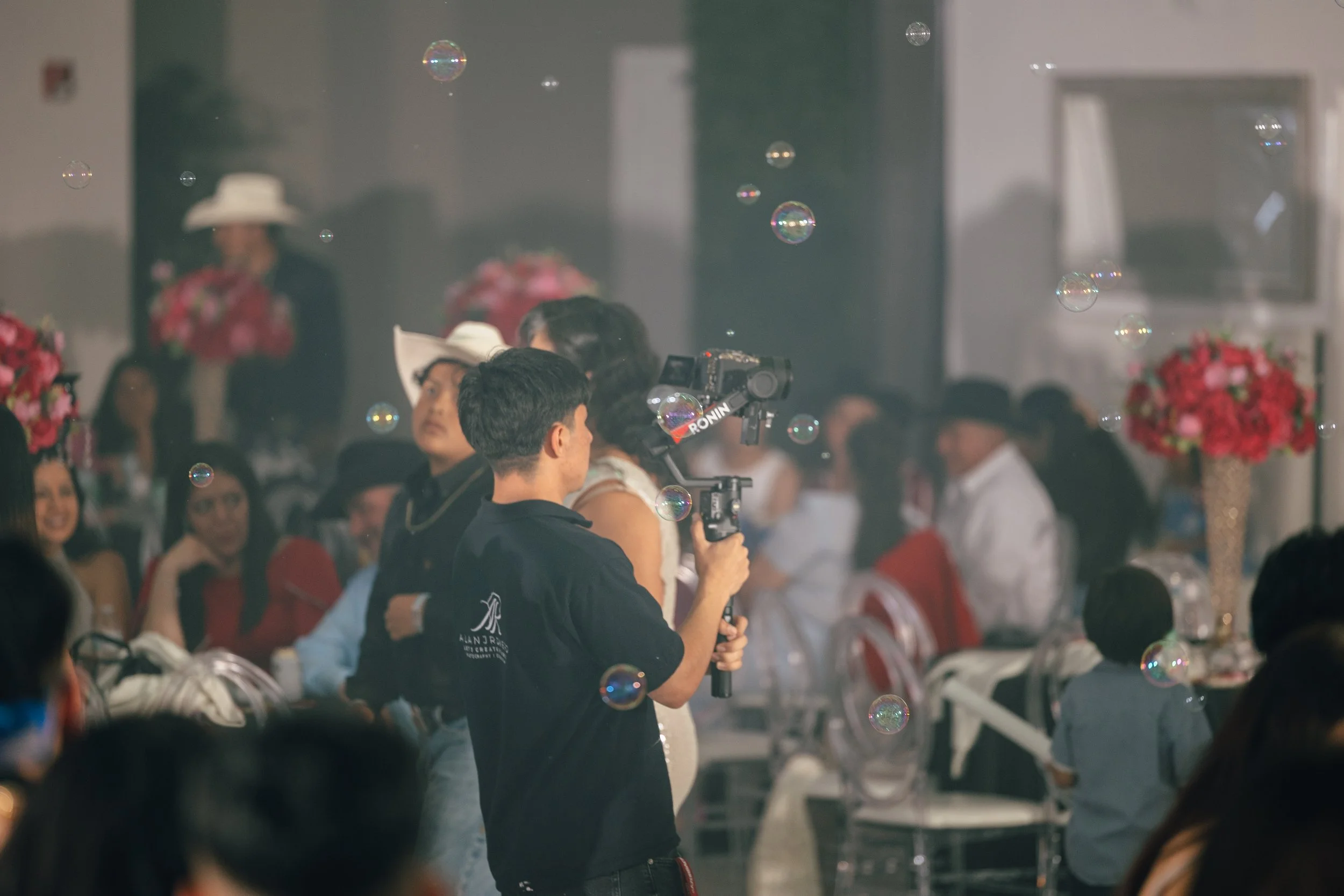 A photographer is taking pictures at a crowded event or celebration, with people dressed up and decorated with pink and red flowers in the background, and soap bubbles floating in the air.