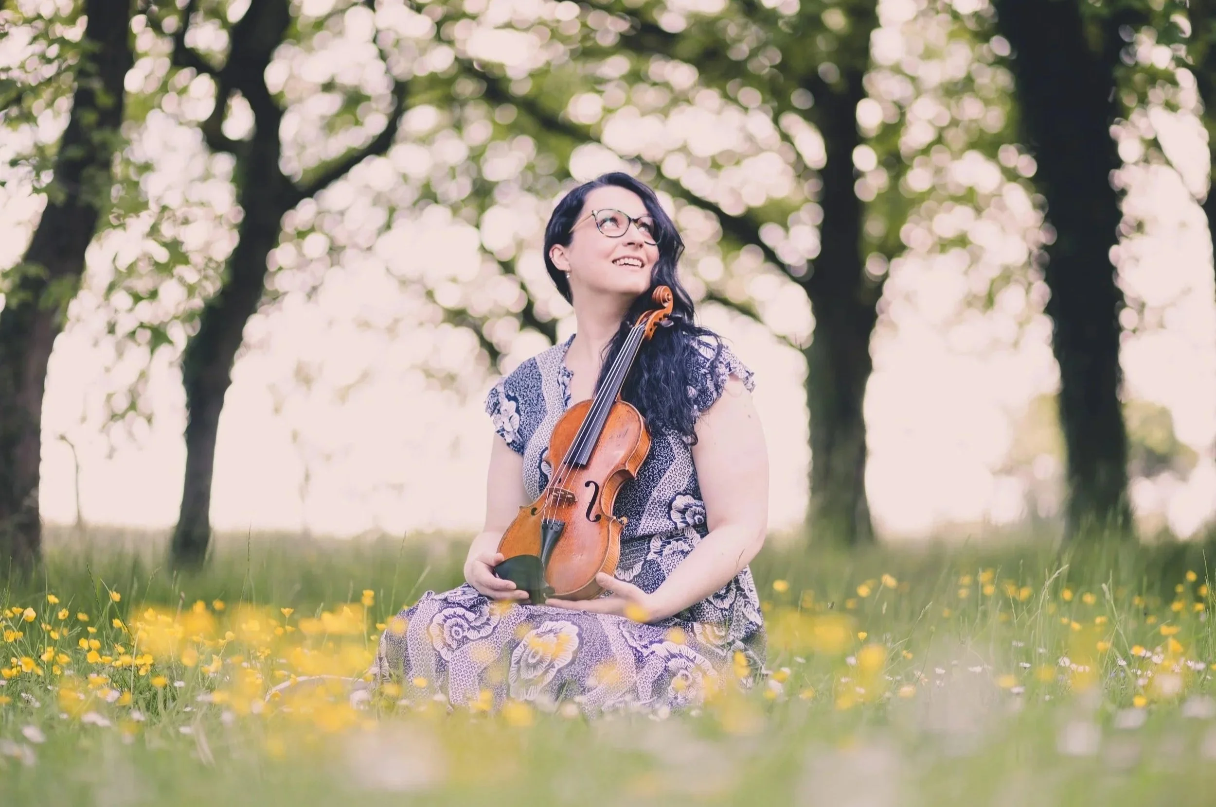 Katy Smith violinist sitting in a field of buttercups holding her violin, with trees in the background.