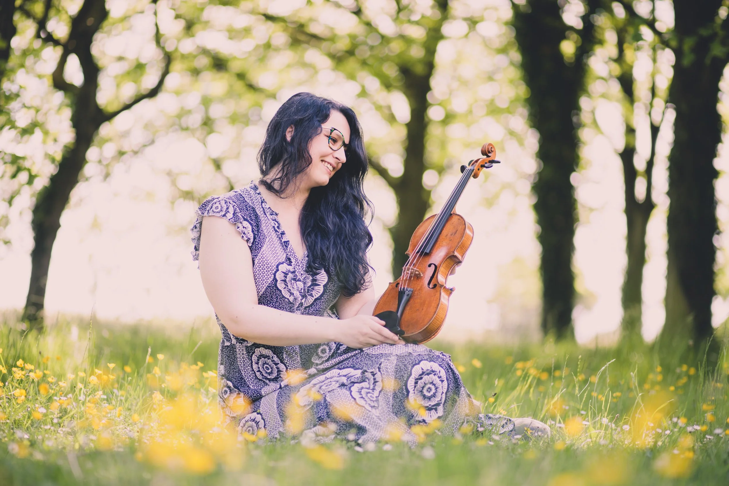 Katy Smith violinist sitting on the grass in a field of buttercups, holding and looking at her violin lovingly amidst trees.
