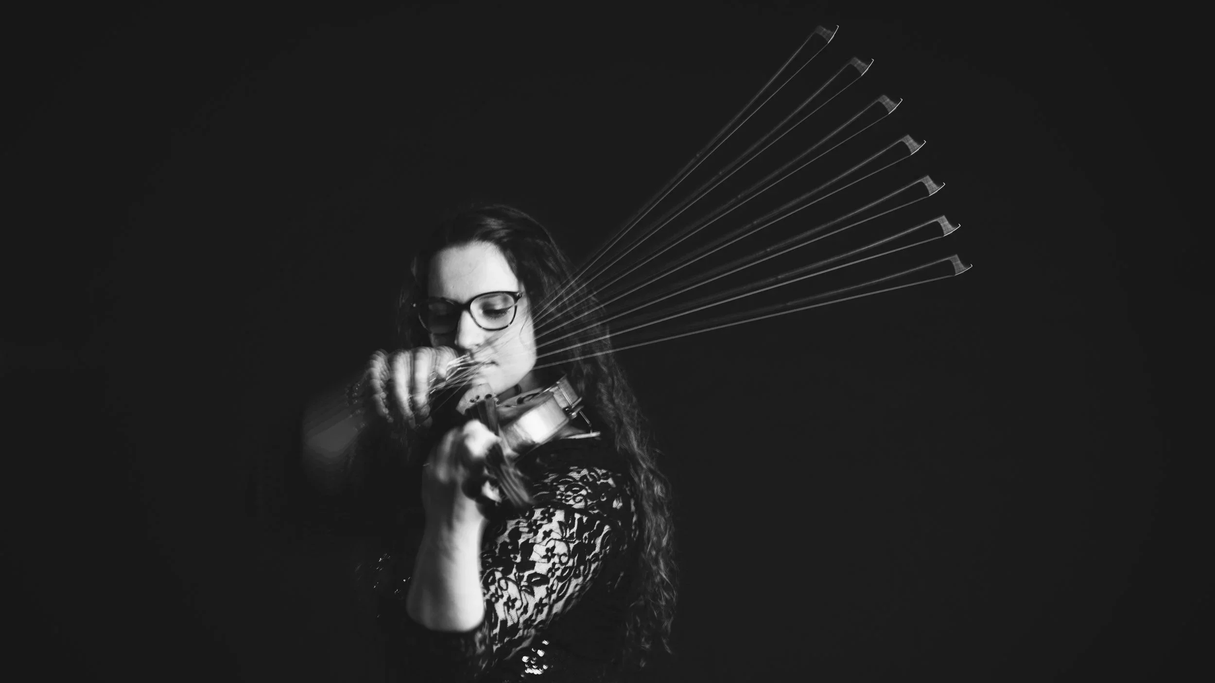 A woman with long curly hair and glasses playing the violin, with the bow in motion, against a dark background.