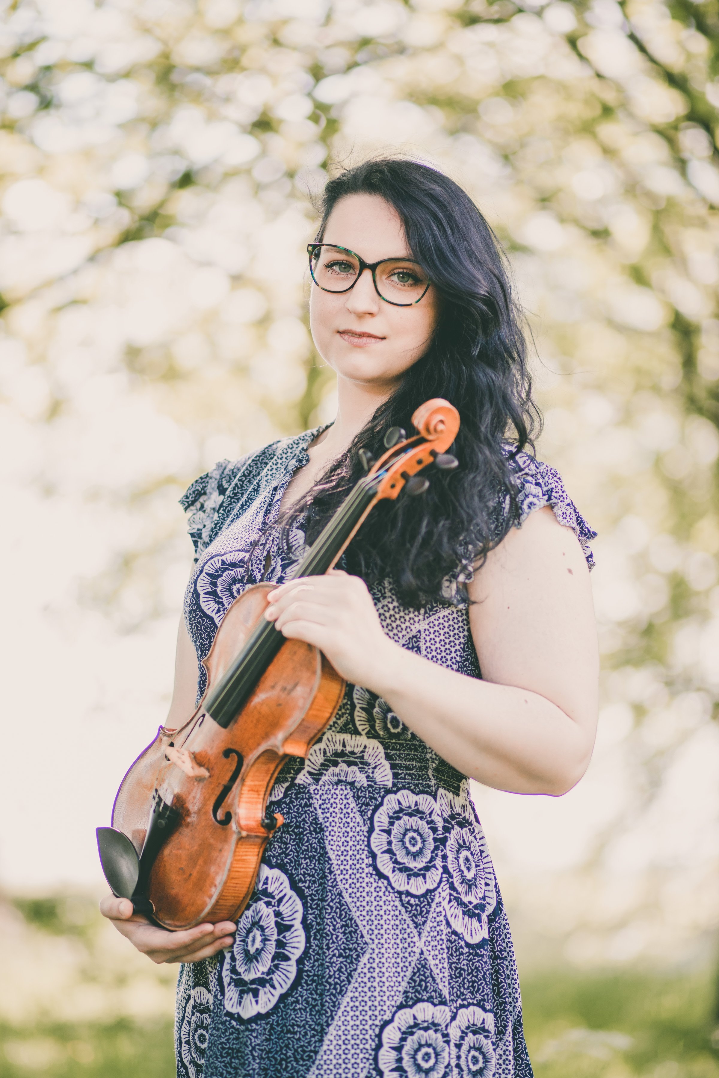 A woman with black wavy hair, glasses, and a patterned dress holds a violin outdoors with trees in the background.