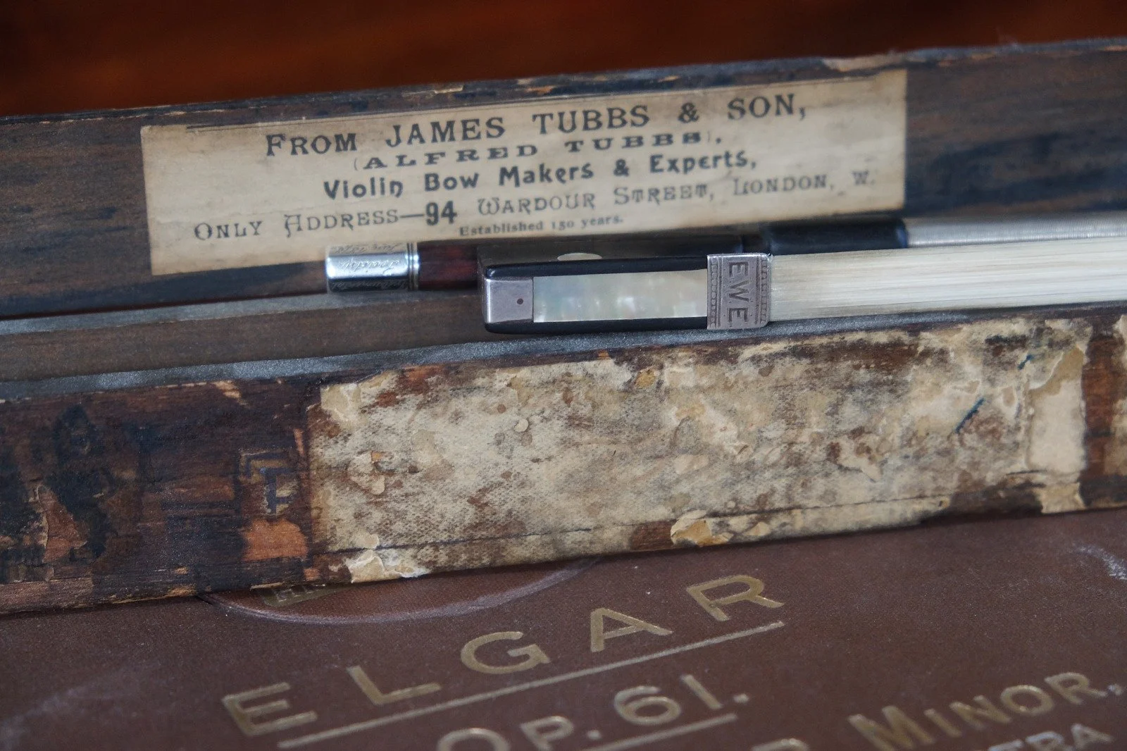 Close-up of a vintage wooden box with a label advertising James Tubbs & Son, violing bow makers in London, and a partially visible handled object inside. The box rests on a surface with the word 'ELECTGAR' visible.