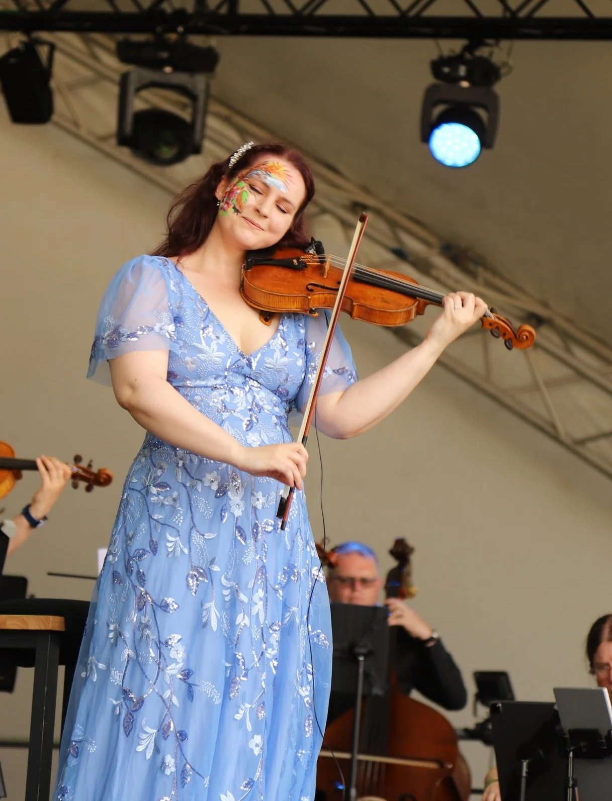 A woman dressed in a light blue, embroidered dress is playing the violin during a musical performance. She has face paint with bright colors and floral designs on her face, and her eyes are closed with a content expression.