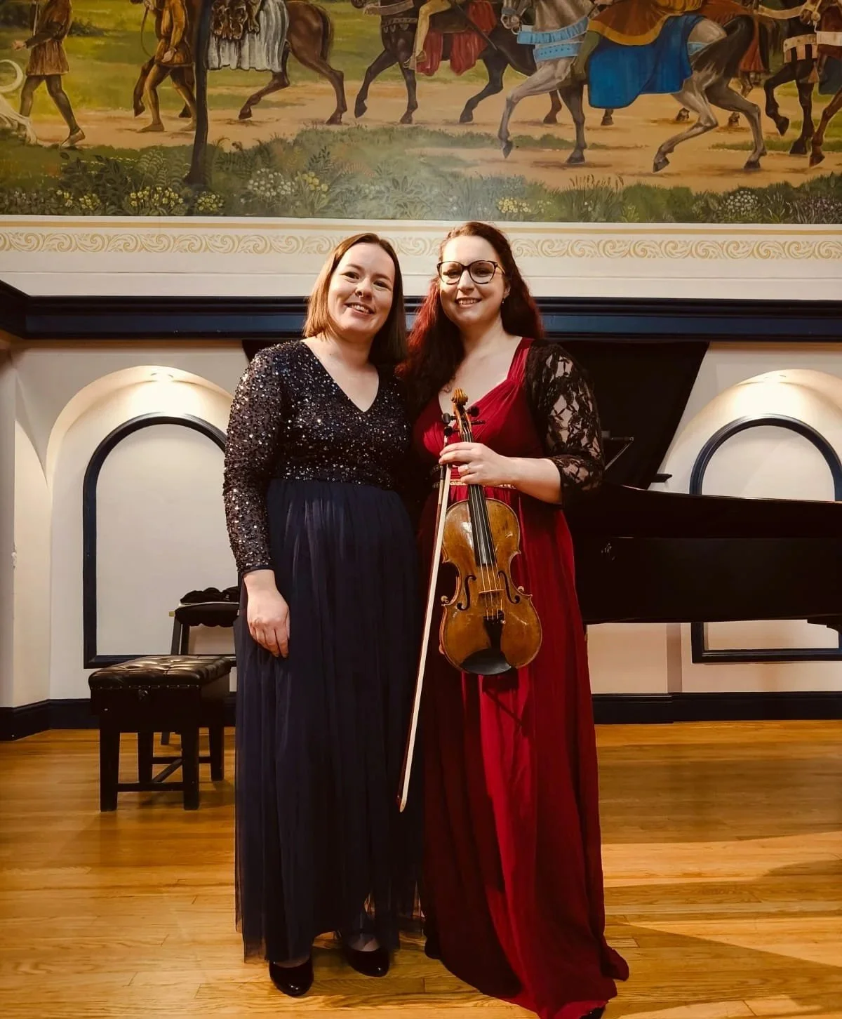 Two women dressed elegantly, one holding a violin, standing on a stage with a grand piano and a mural of historic scenes in the background.