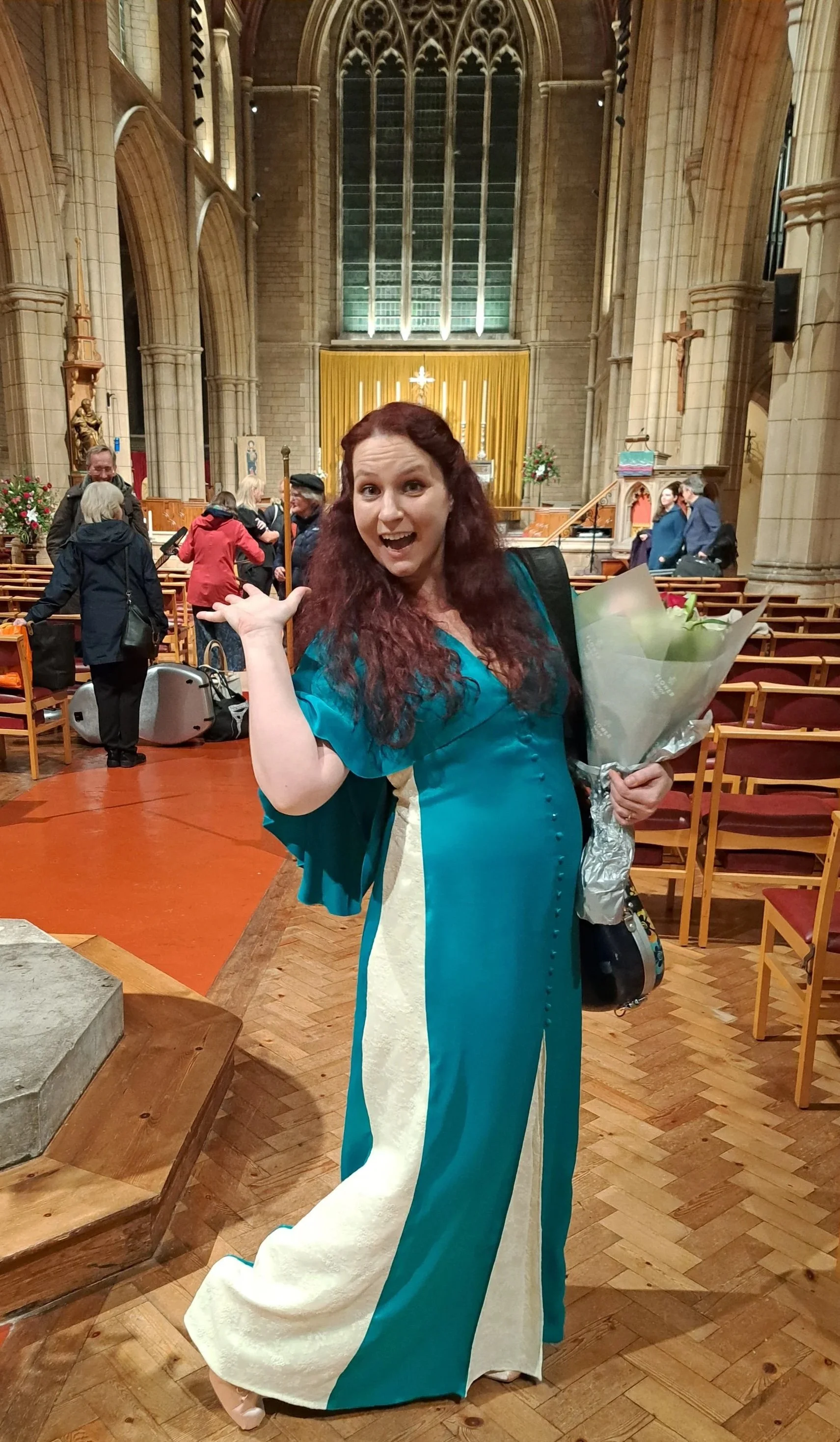 A woman with long red hair wearing a teal and beige gown, holding a bouquet of flowers, standing inside a church with an enthusiastic expression, surrounded by other people and church decor.