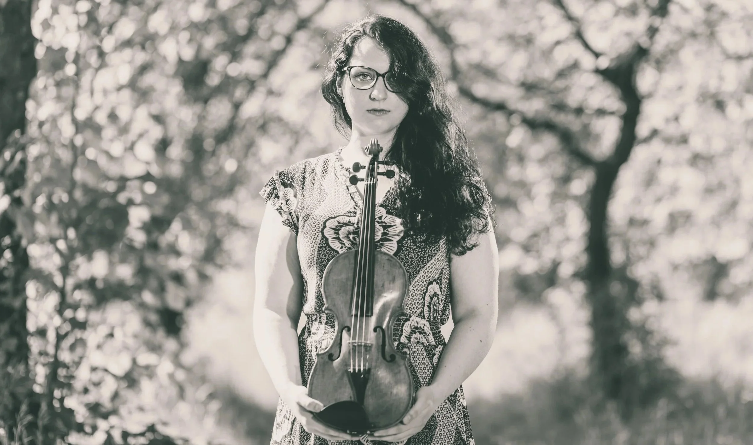 Black and white photo of a woman with long dark curly hair, glasses, wearing a patterned dress, holding a violin in front of her chest, outdoors with trees in the background.