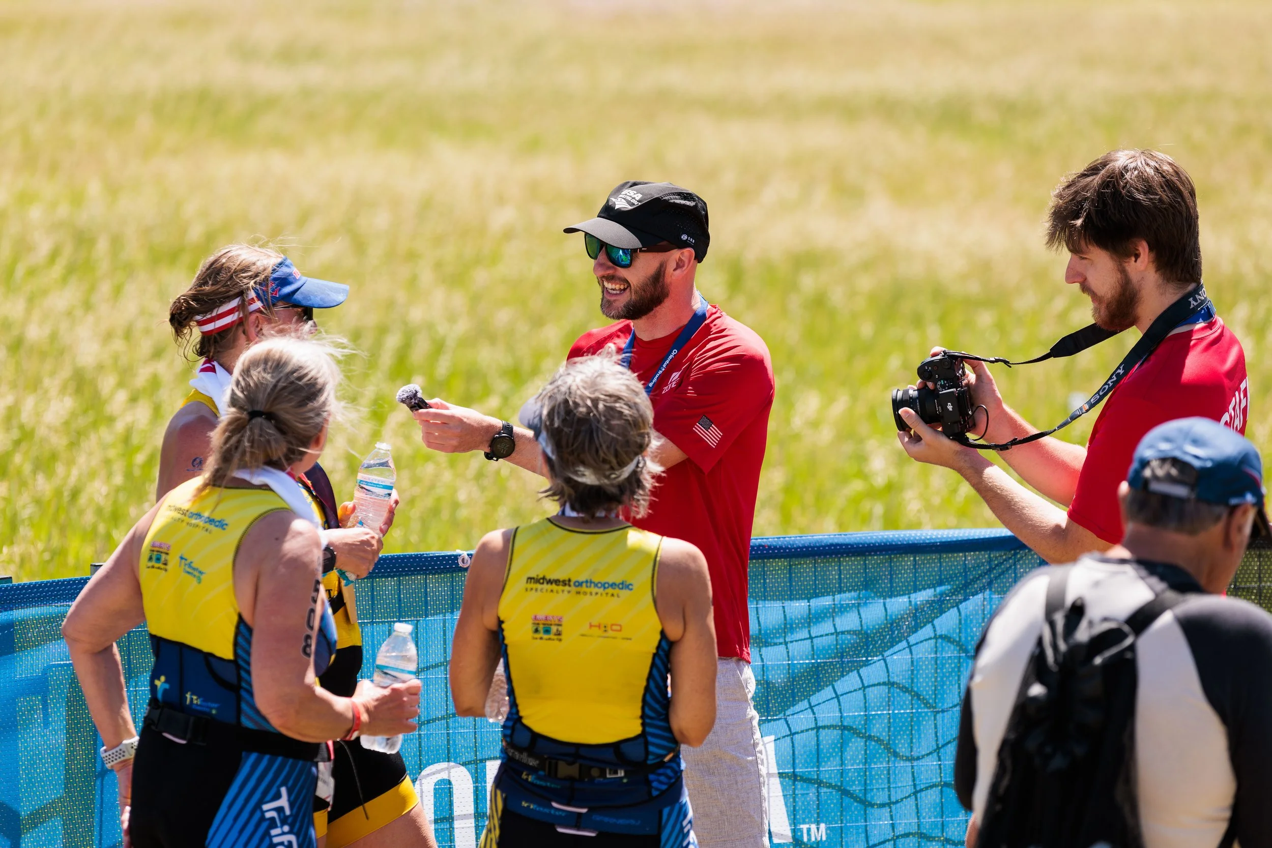 Group of athletes in yellow and black uniforms being interviewed by a man in a red shirt and black cap, with another man in a red shirt filming them, outdoors in a grassy field.
