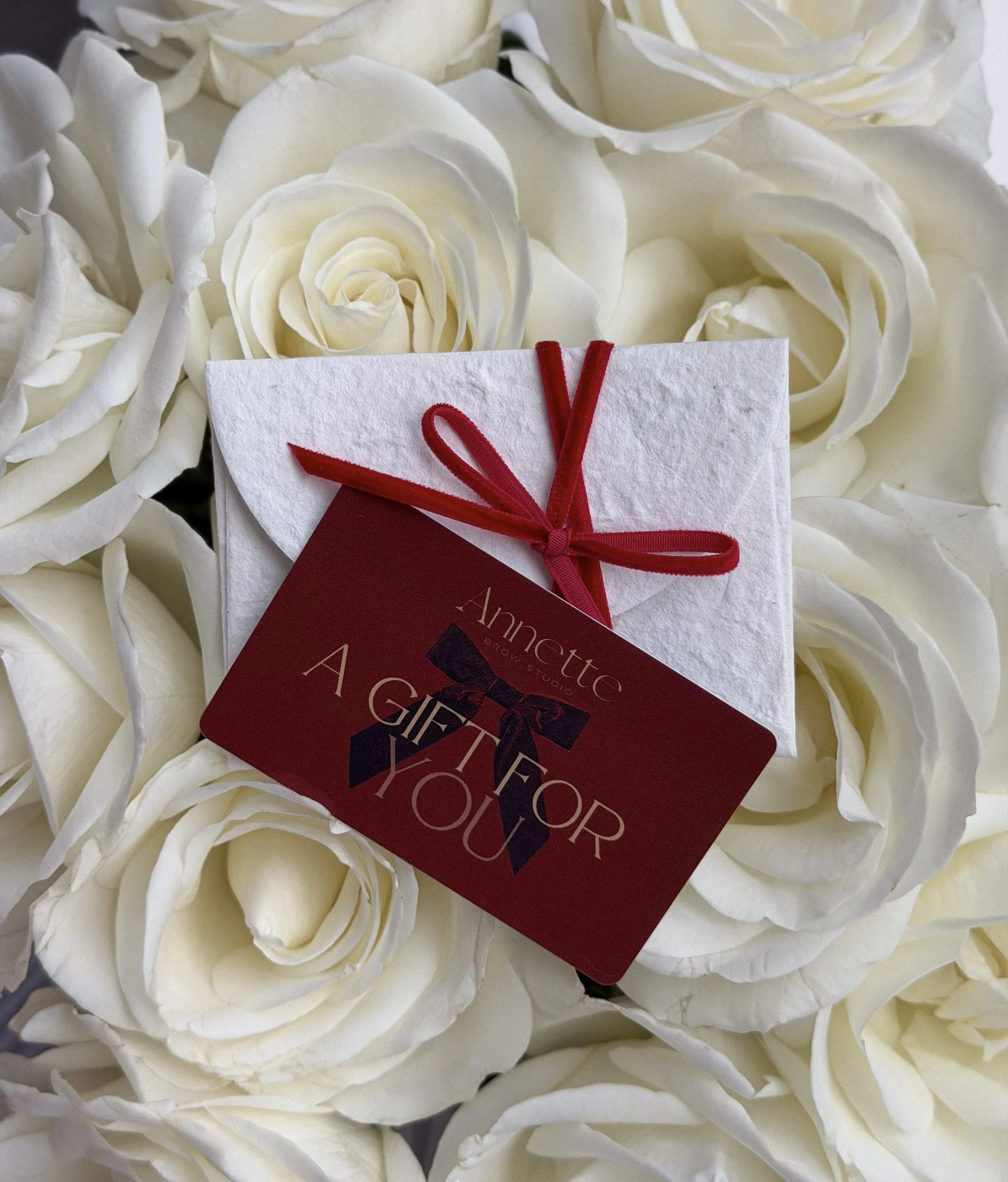 A close-up of white roses with a small gift card and red ribbon on top, surrounded by white petals.
