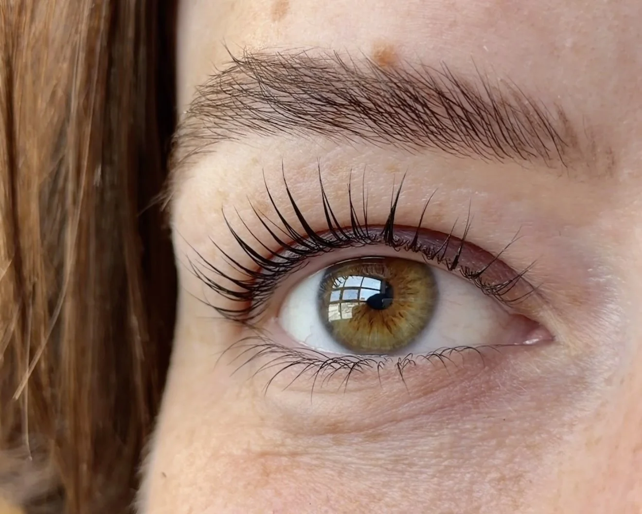 Close-up of a person's eye, showing brown iris, long eyelashes, and a reflection of a window in the eye.