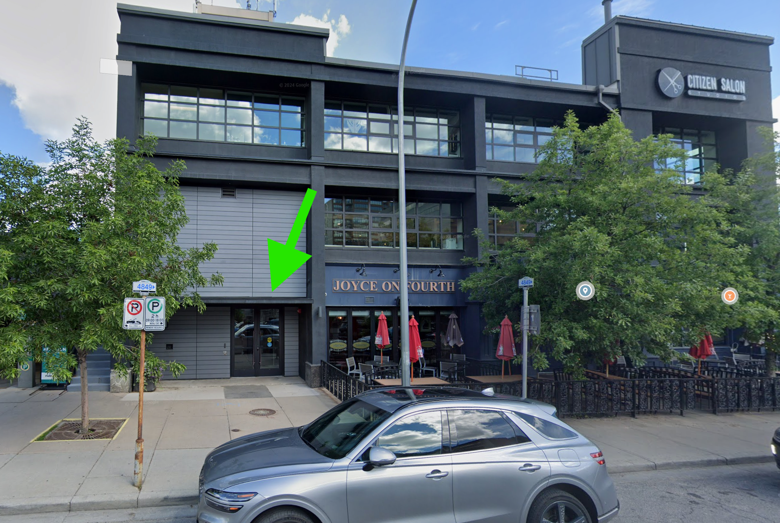 Exterior view of a multi-story building with a sign that reads "Joyce on Fourth," with outdoor patio seating and umbrellas, two trees, street parking, and a no parking sign.